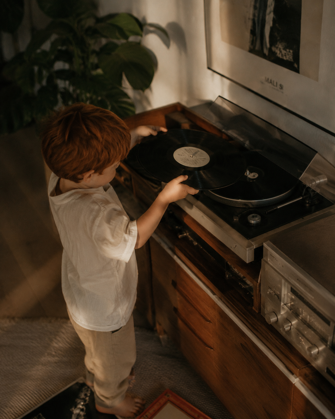 Enfant roux de dos actionnant une platine vinyle – salon, lumière chaude et plante verte