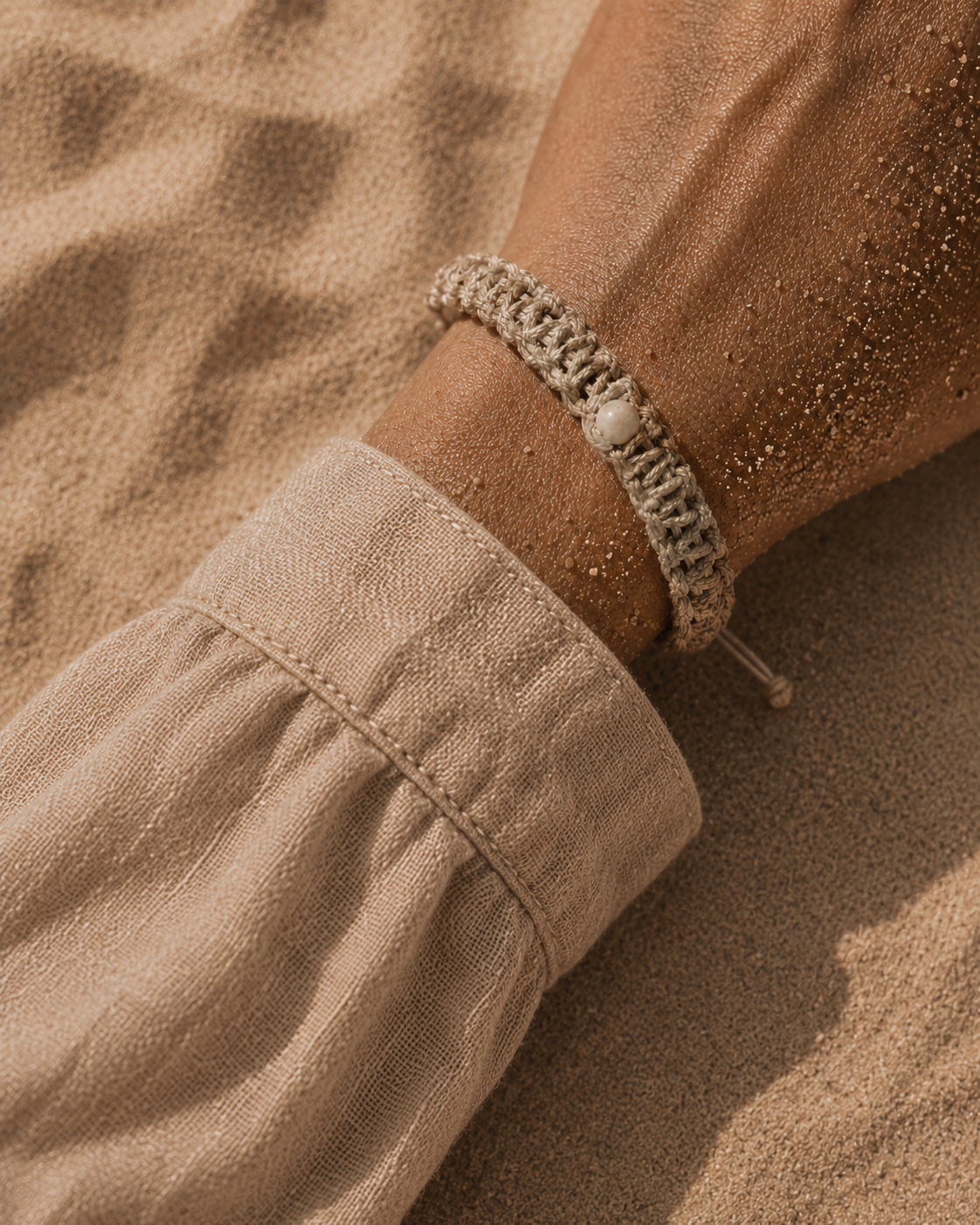 Gros plan poignet avec bracelet macramé beige – perle blanche, sable doré sur peau, manche lin