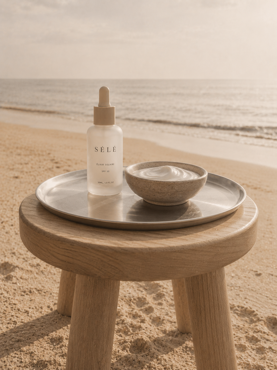 Flacon sérum et bol crème sur plateau métal – tabouret bois sur plage, horizon mer, coucher de soleil