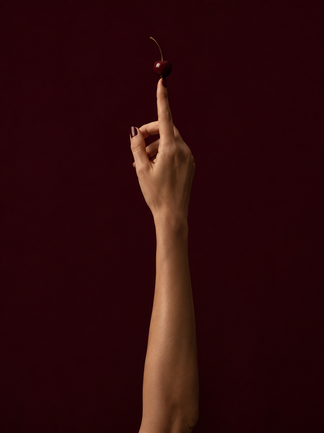 Bras et main féminine tendus vers le haut, index soutenant une cerise rouge sur fond bordeaux profond, ongles bordeaux