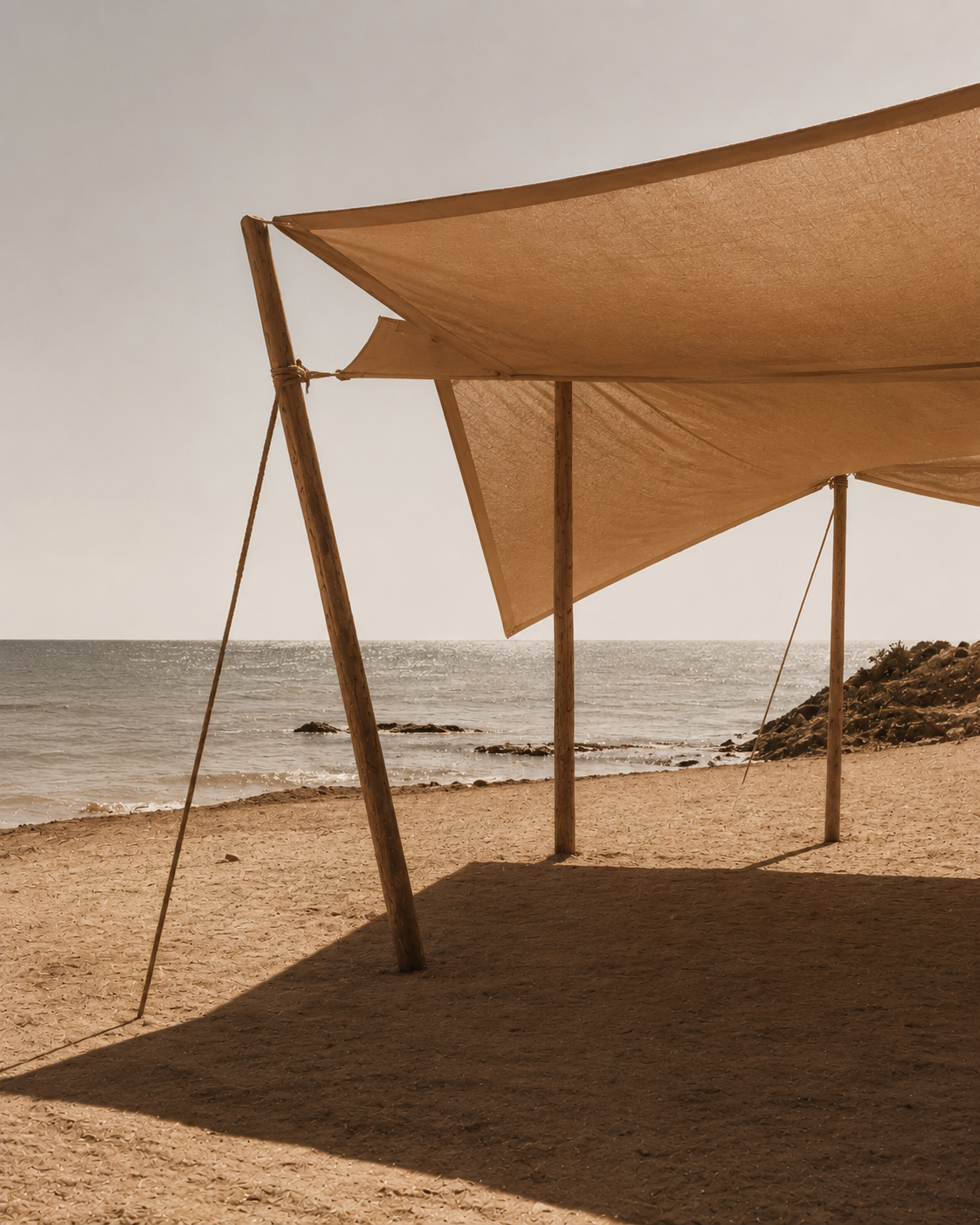 Voile de lin beige sur poteaux bambou en bord de plage – mer, rochers, ciel terne, ombre portée