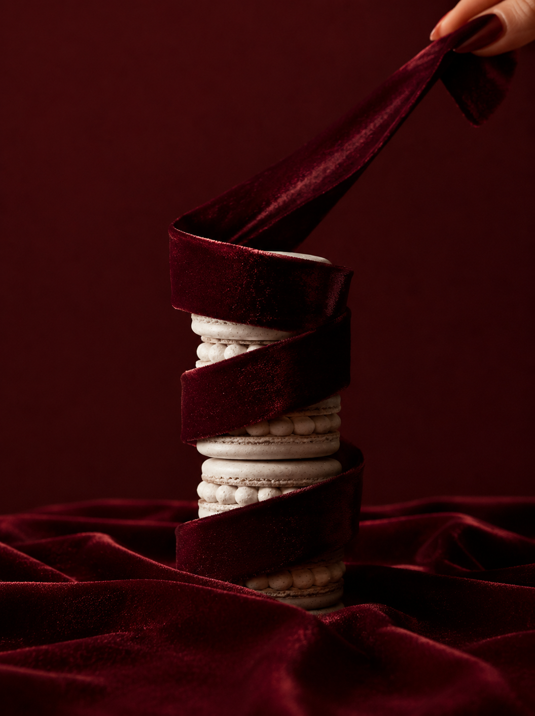 Tour de macarons blancs serrée enroulée d'un ruban de velours bordeaux déroulé vers le haut par une main, drapé bordeaux en fond