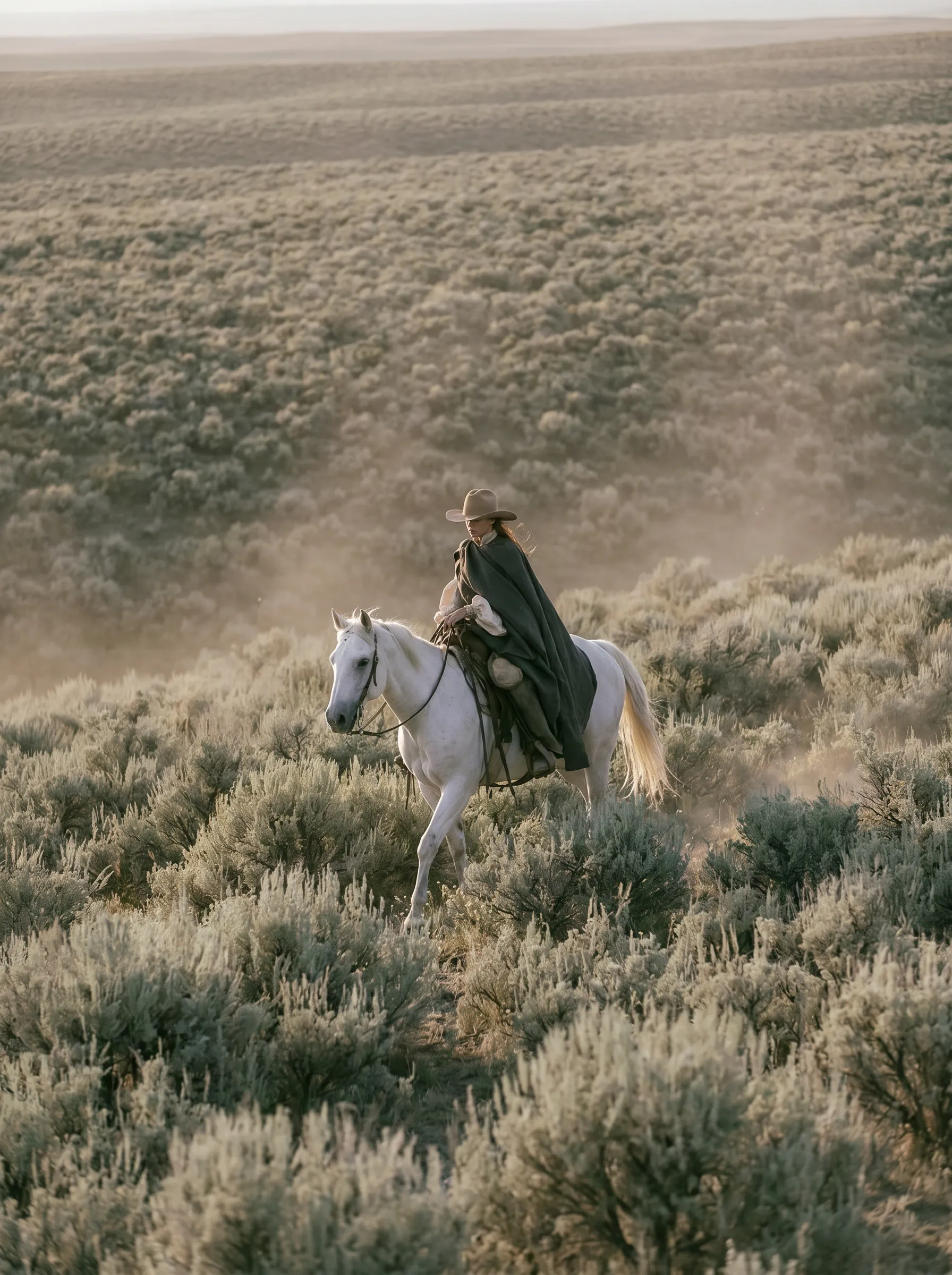 Cavalière en cape noire et chapeau à cheval blanc traversant une steppe broussailleuse de sagebrush