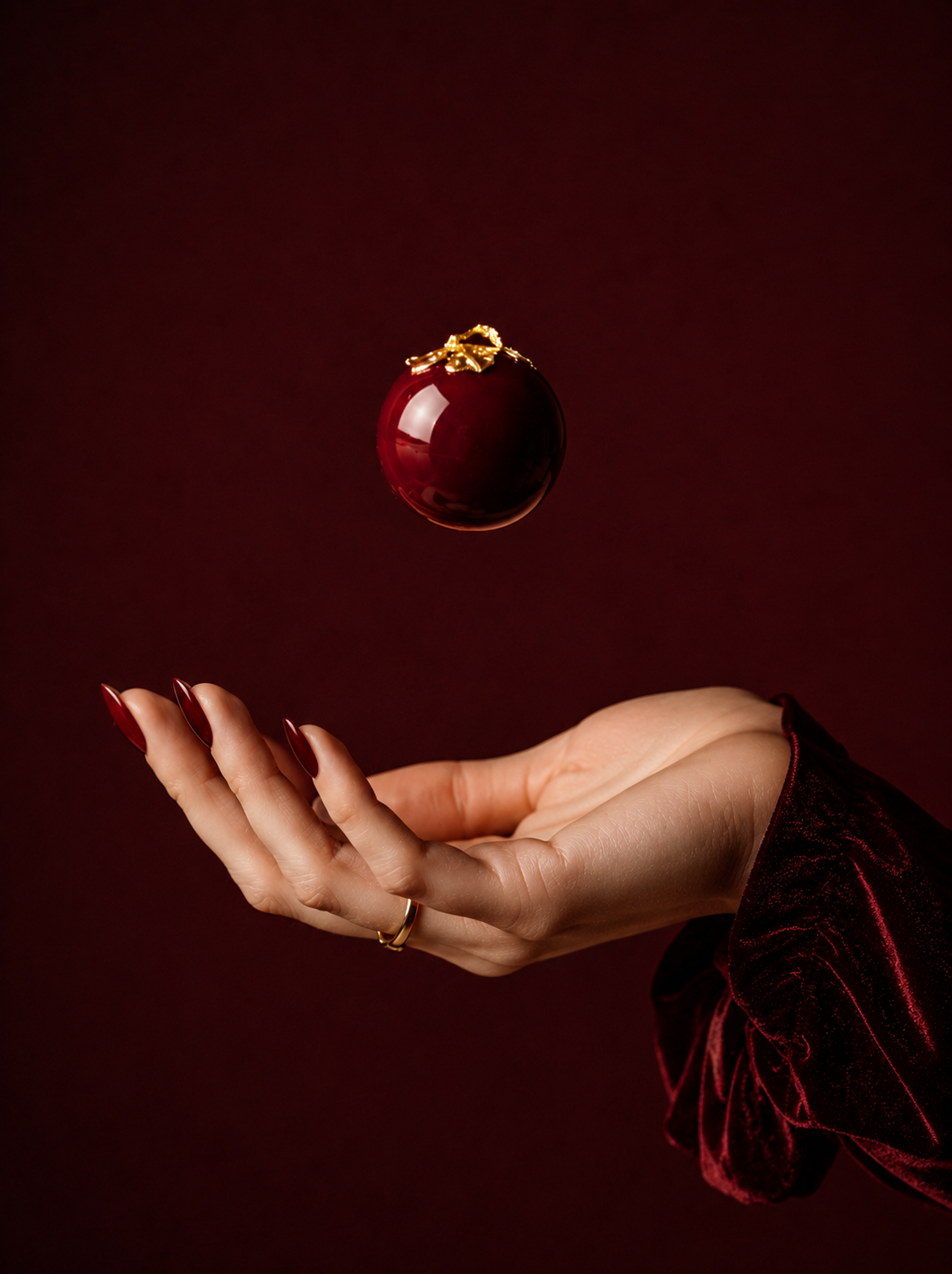 Main féminine avec bague dorée, entremets sphérique bordeaux en lévitation – fond bordeaux