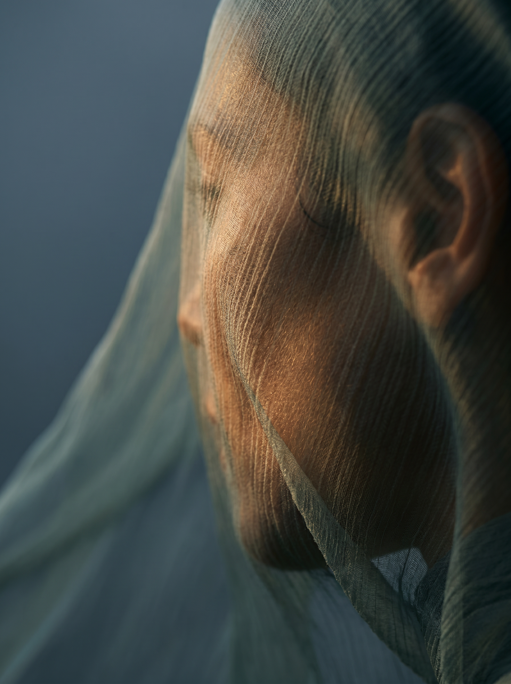 Portrait gros plan femme à travers voile gaze – lumière chaude dorée sur le visage, fond bleu-gris