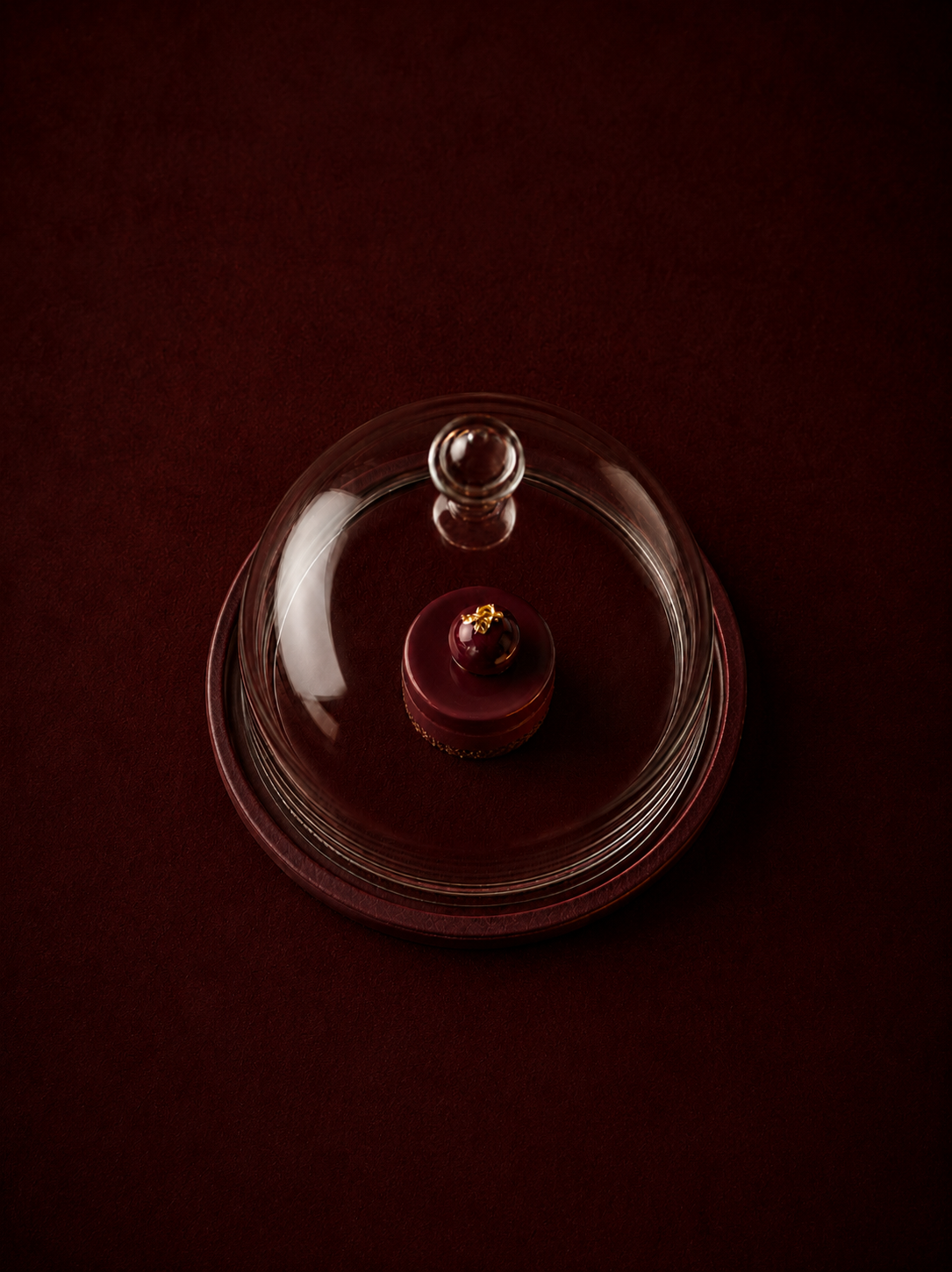 Vue aérienne sur petite pâtisserie rouge individuelle sous cloche en verre transparente, fond bordeaux profond, composition minimaliste