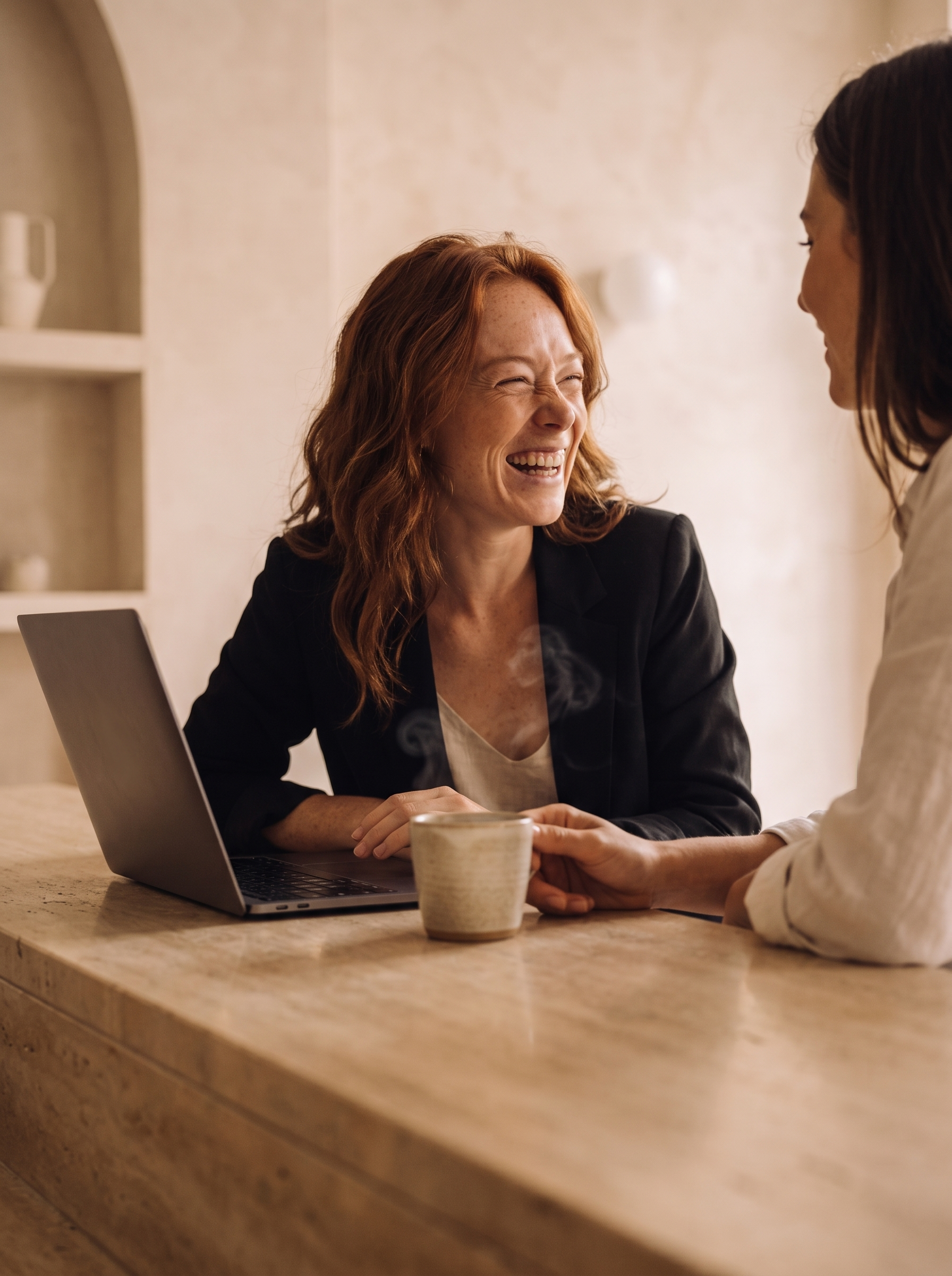 Femme rousse en blazer noir riant avec une collègue devant son laptop dans un café