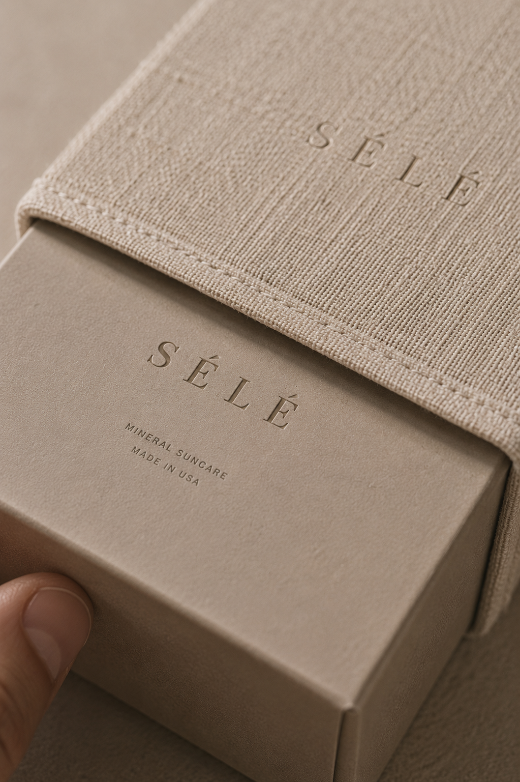 Main extrayant une boîte kraft d'un coffret tissu texturé beige – packaging luxe, fond neutre