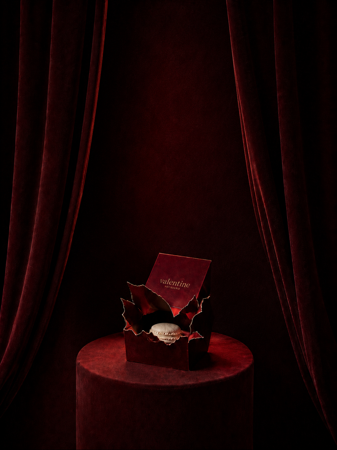 Boîte Valentine ouverte avec macaron blanc posé dessus et feuilles de houx dorées sur présentoir velours rouge bordeaux, drapé bordeaux