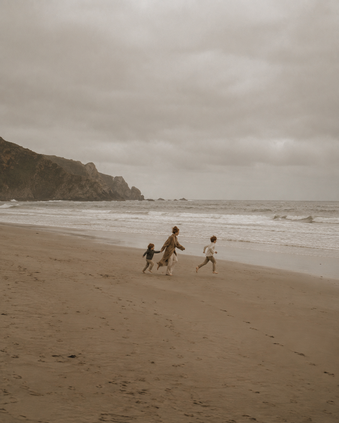 Femme et deux enfants courant sur une plage de sable – ciel nuageux, mer et falaises