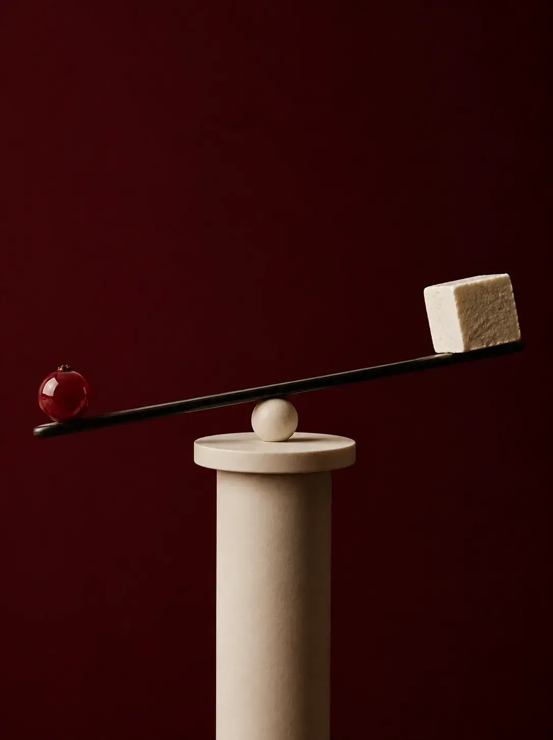 Balance minimaliste sur colonne travertin – sphère rouge et cube beige en équilibre, fond bordeaux