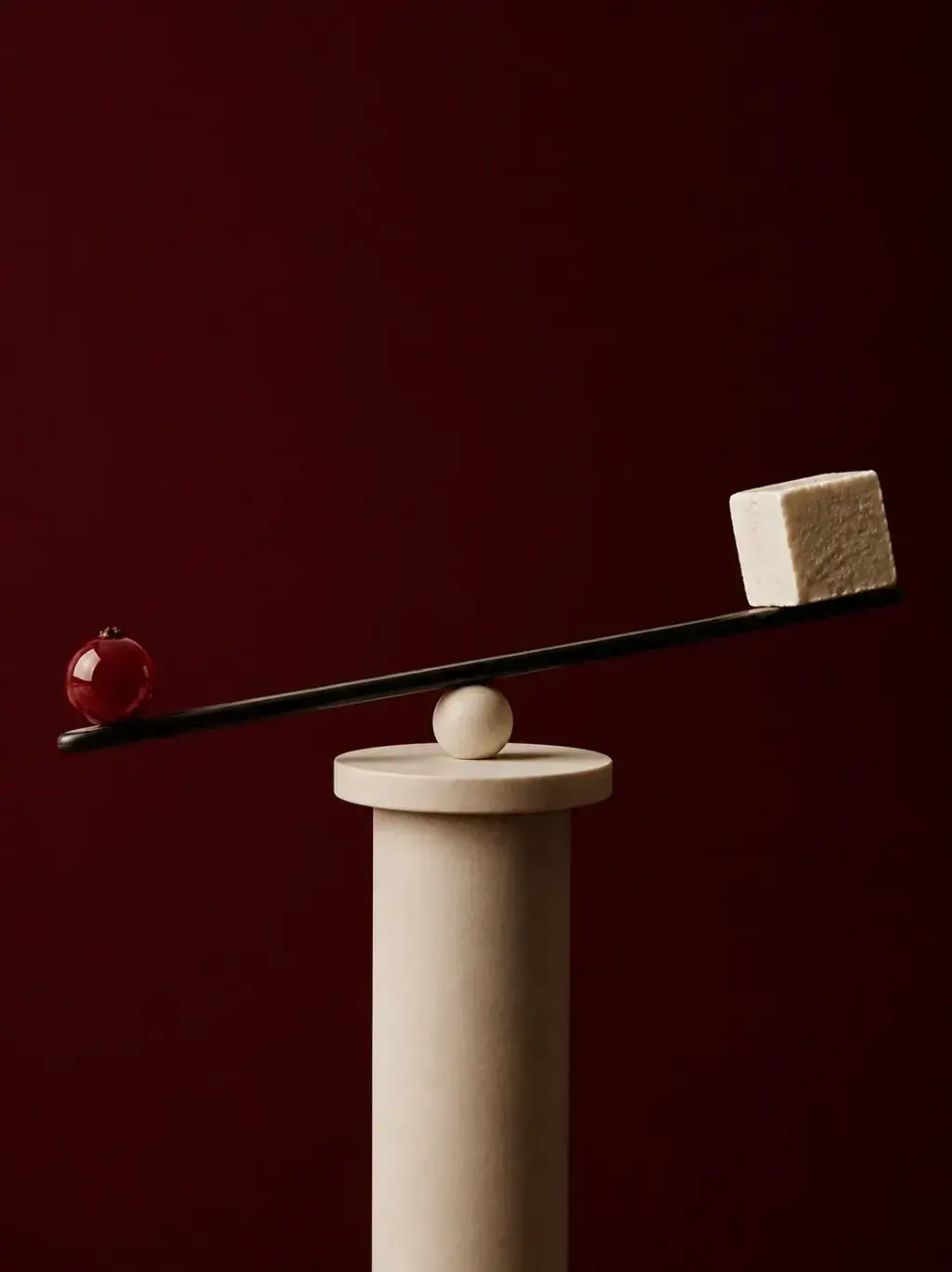 Balance minimaliste sur colonne travertin – sphère rouge et cube beige en équilibre, fond bordeaux