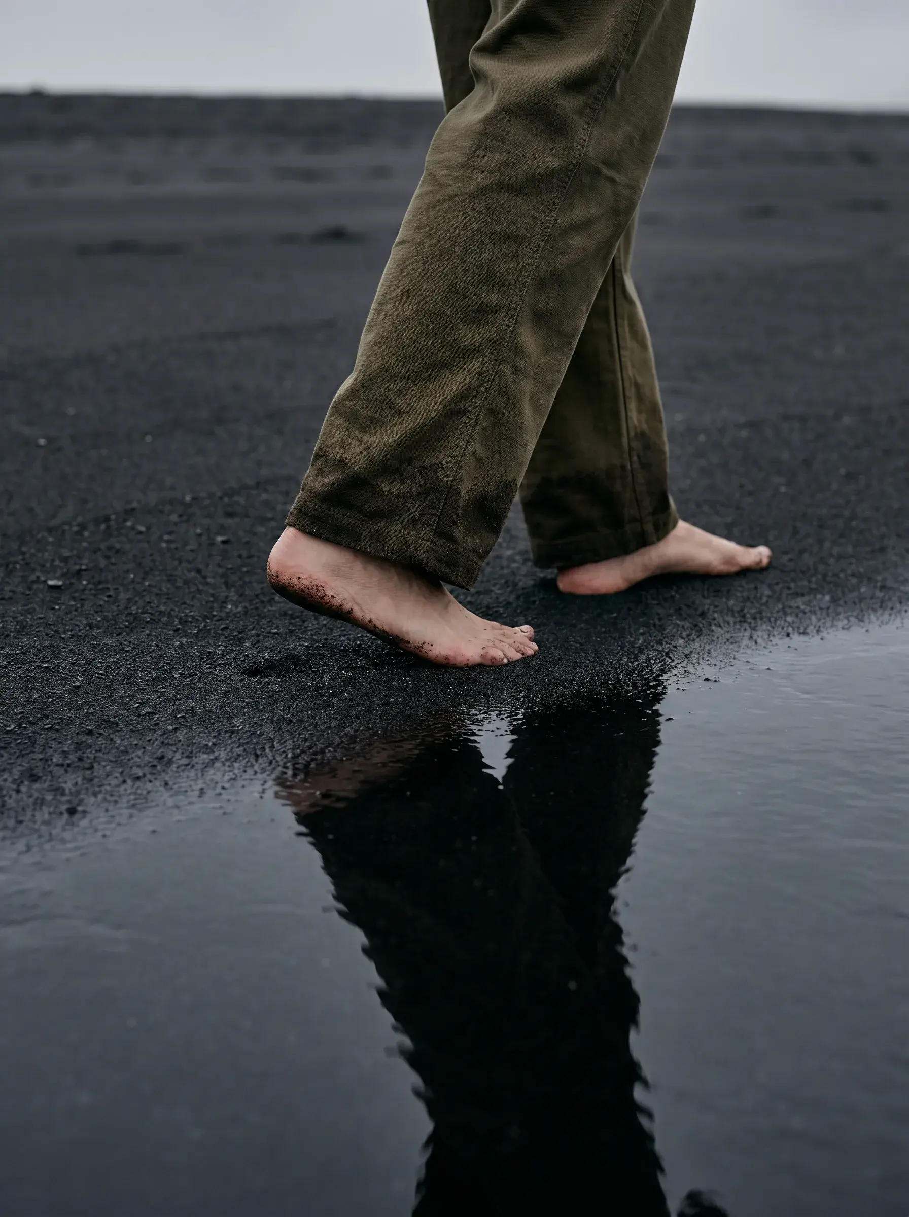 Bas de pantalon cargo kaki et pieds nus sur sable volcanique noir mouillé