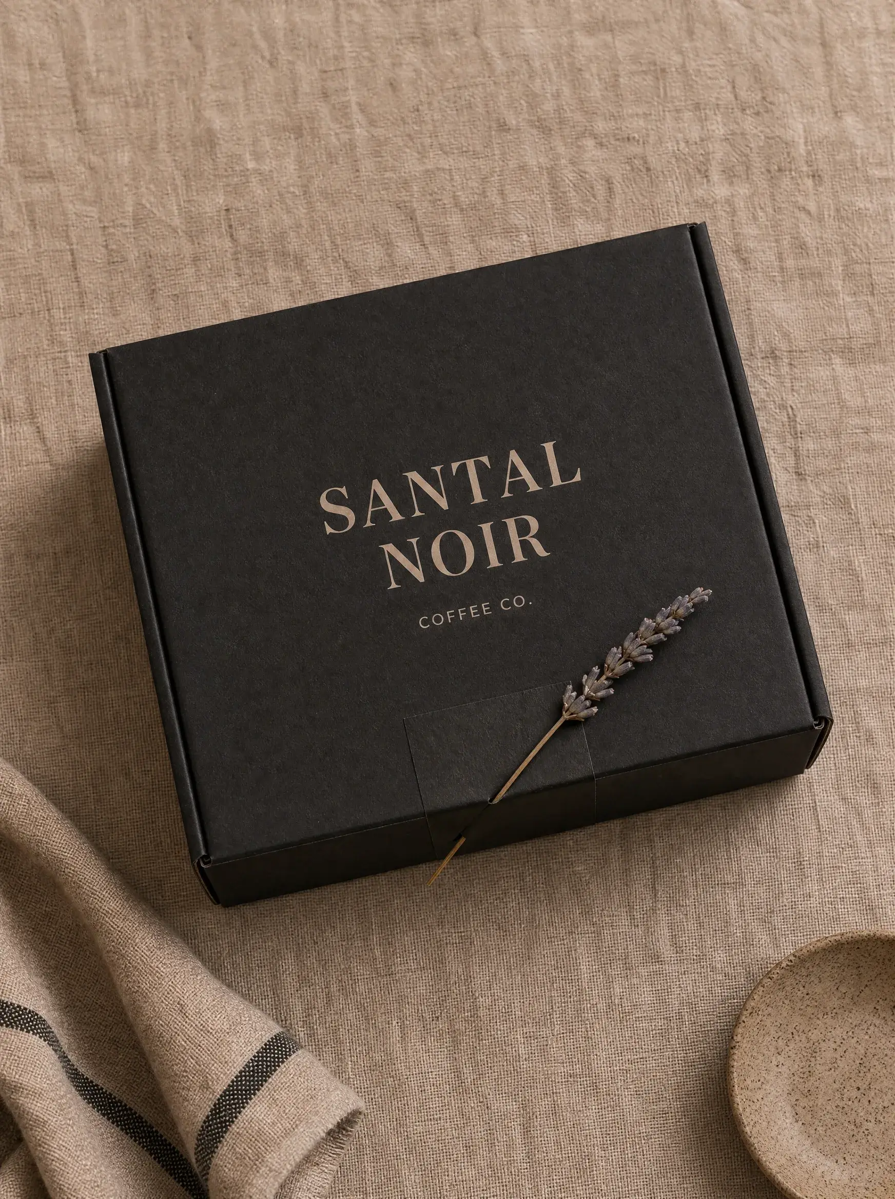 Boîte cadeau noire fermée avec branche lavande – nappe lin rayée, cuillère bois, composition épurée
