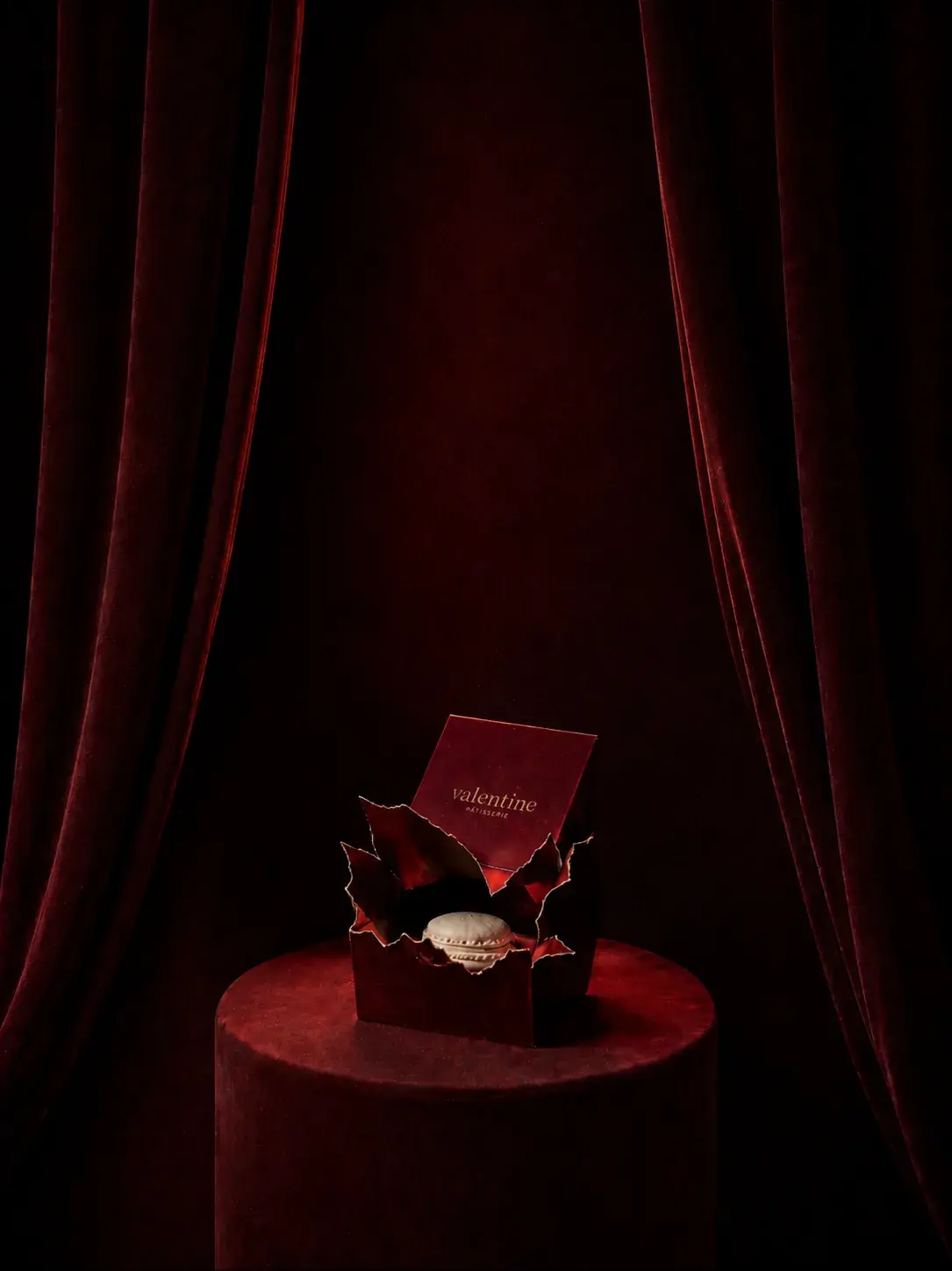 Boîte Valentine ouverte avec macaron blanc posé dessus et feuilles de houx dorées sur présentoir velours rouge bordeaux, drapé bordeaux