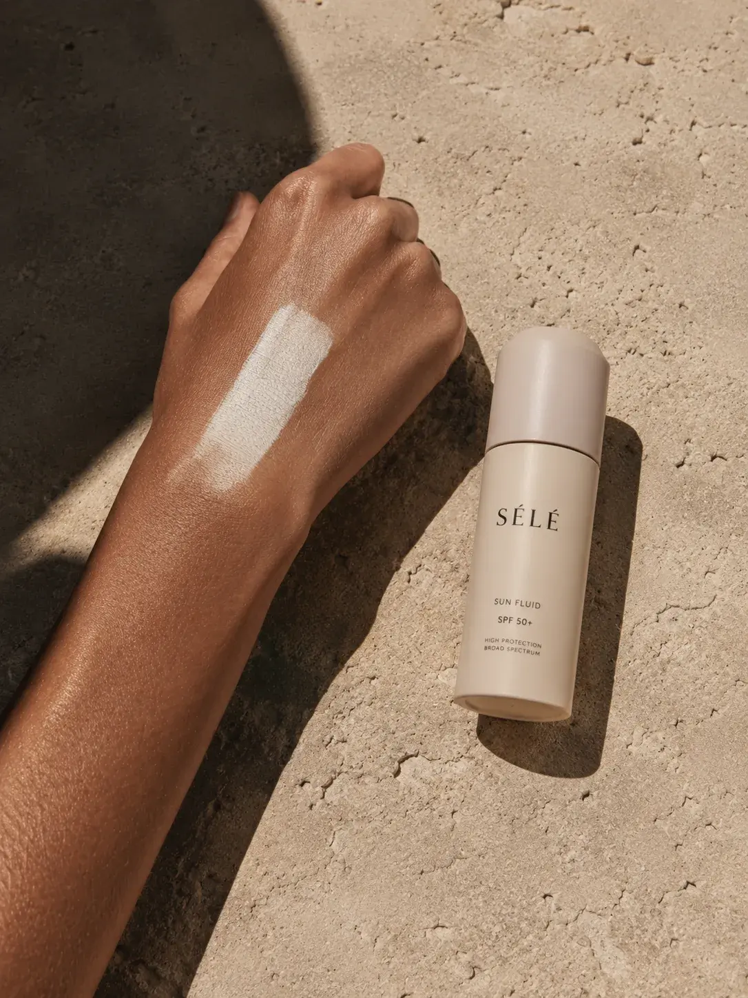 Bras bronzé avec trace de fluide solaire – flacon beige posé sur sable, ombre portée
