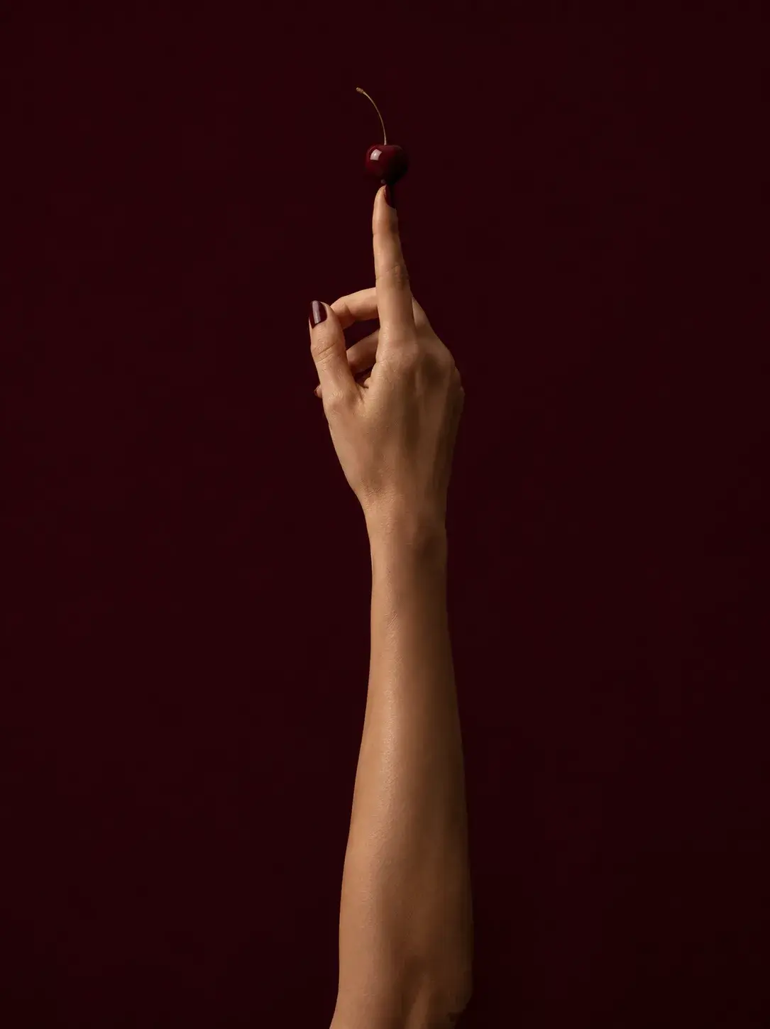 Bras et main féminine tendus vers le haut, index soutenant une cerise rouge sur fond bordeaux profond, ongles bordeaux