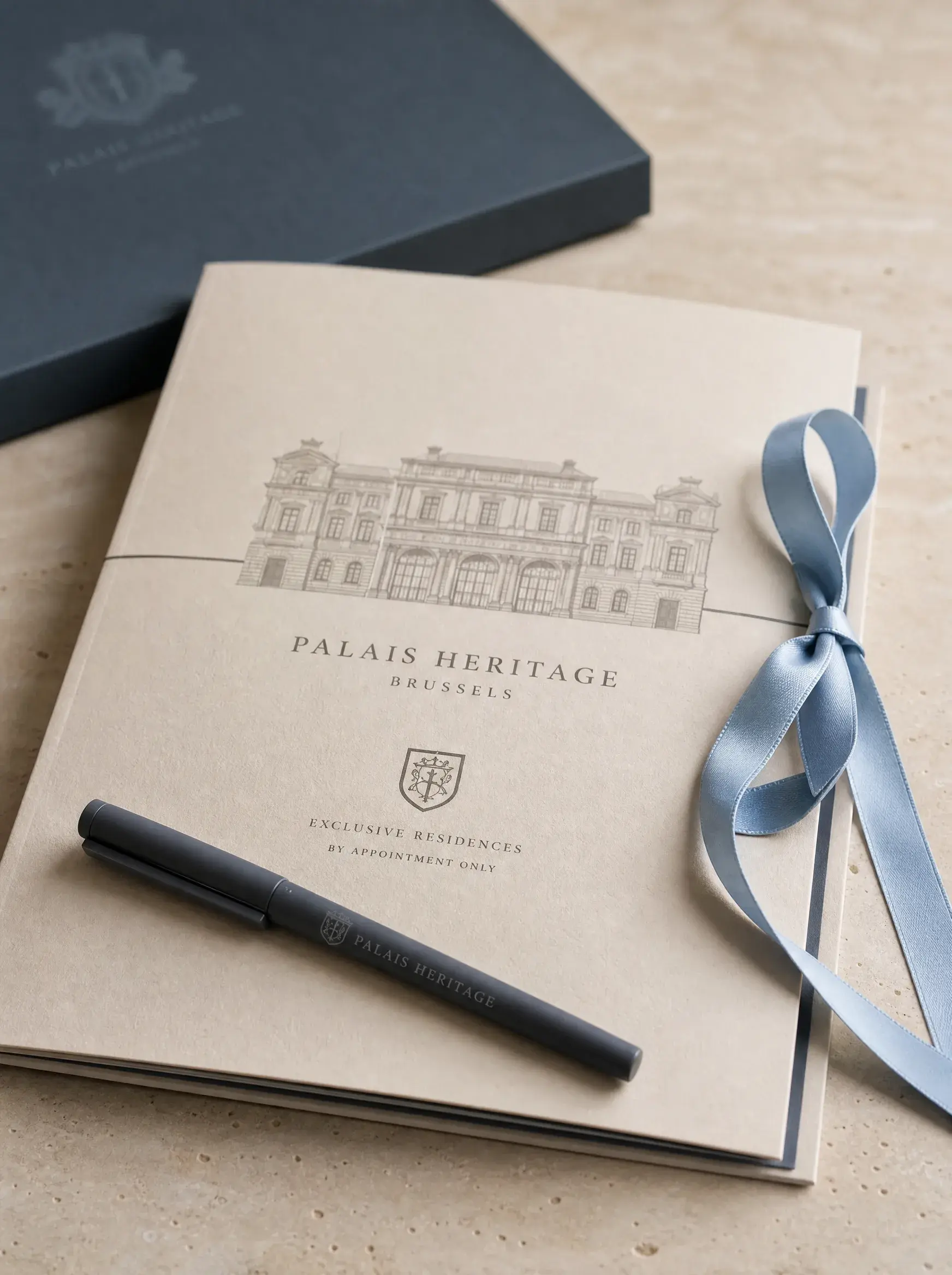 Brochure de prestige Palais Heritage Brussels avec stylo noir et ruban bleu sur fond pierre