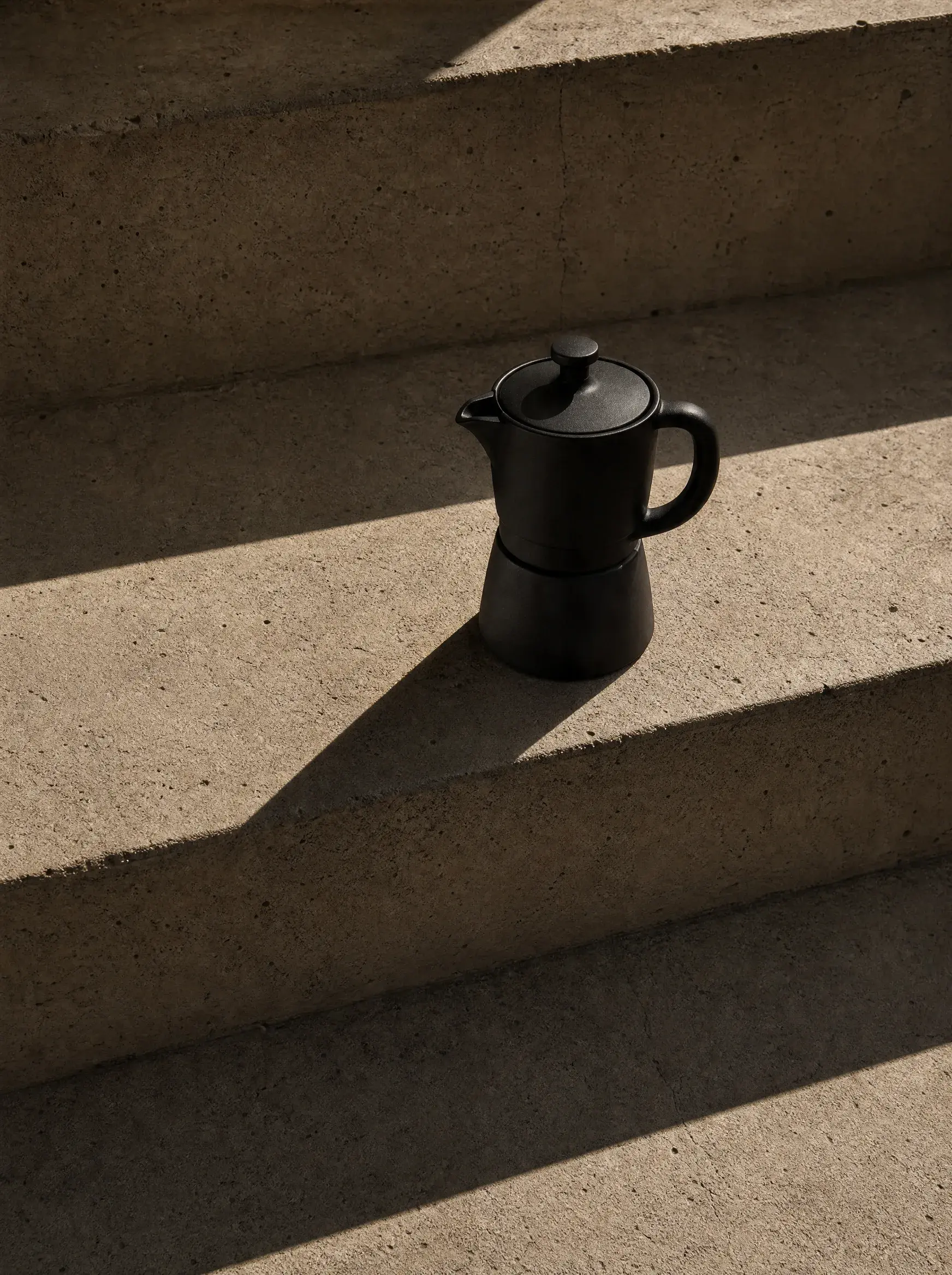 Cafetière moka noire sur marches béton – ombres portées, lumière directionnelle, minimalisme