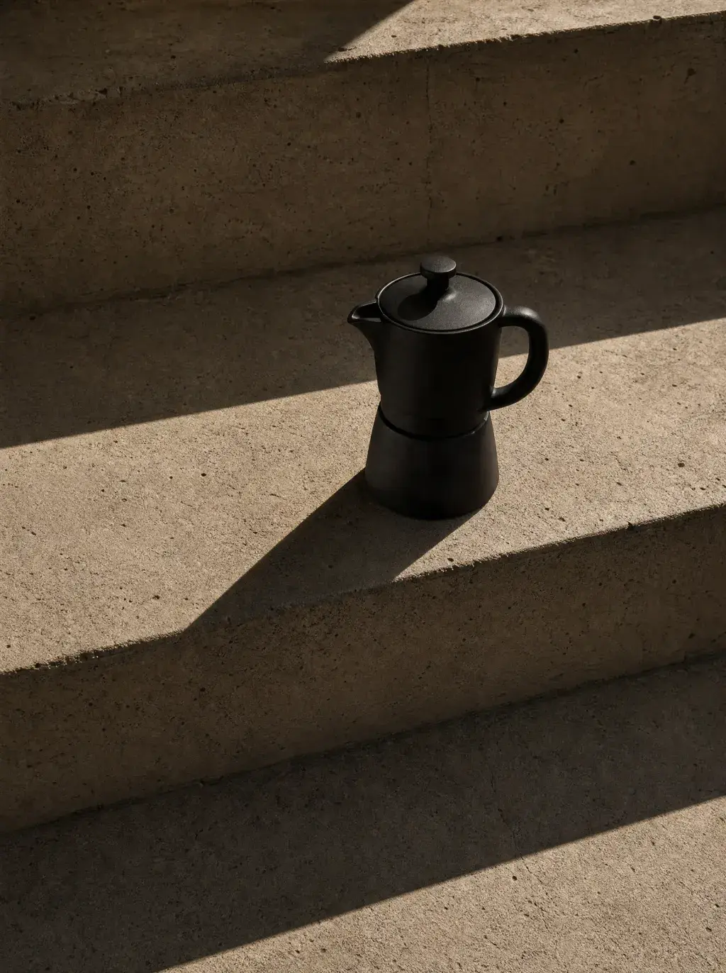 Cafetière moka noire sur marches béton – ombres portées, lumière directionnelle, minimalisme