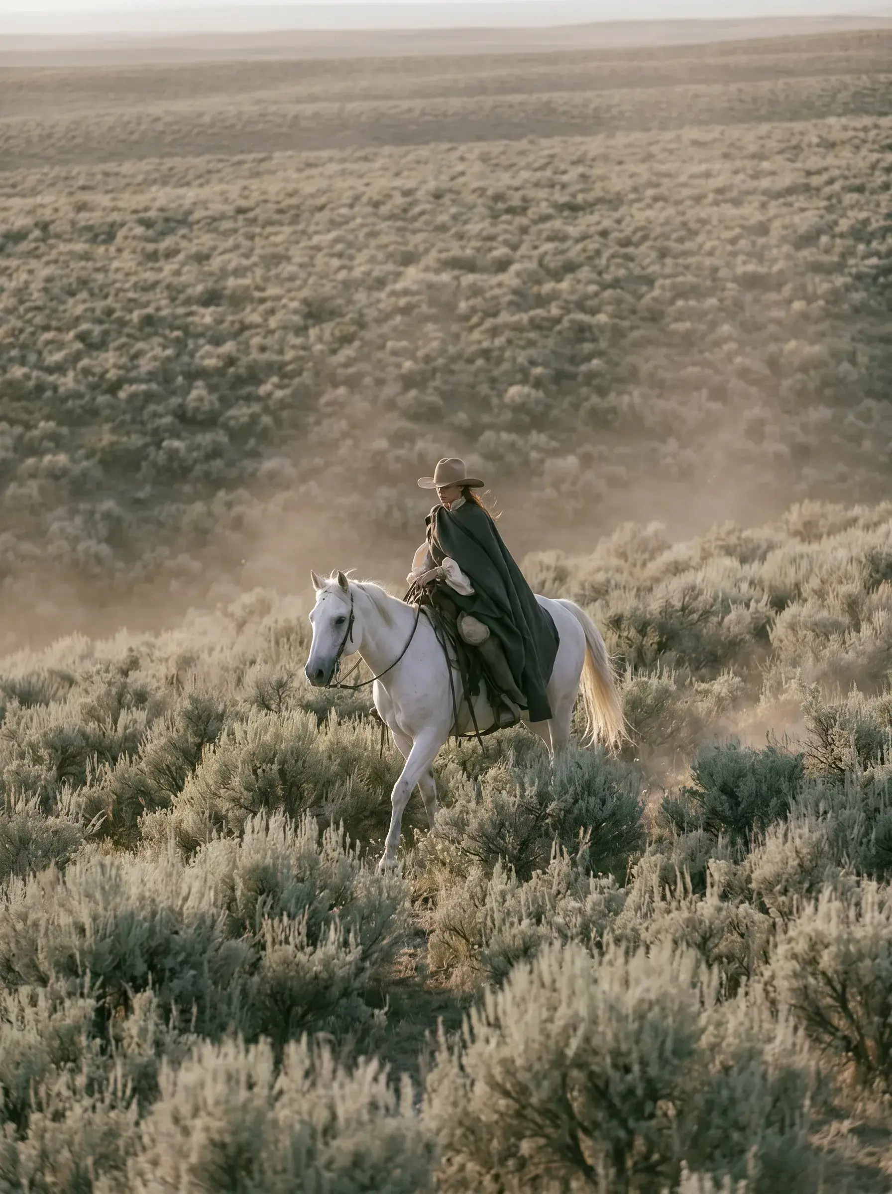 Cavalière en cape noire et chapeau à cheval blanc traversant une steppe broussailleuse de sagebrush