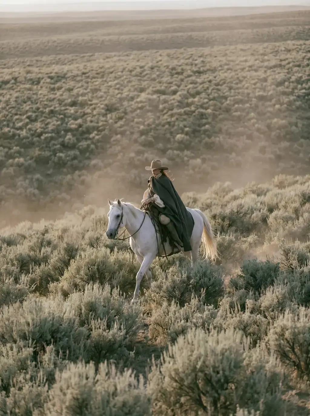 Cavalière en cape noire et chapeau à cheval blanc traversant une steppe broussailleuse de sagebrush