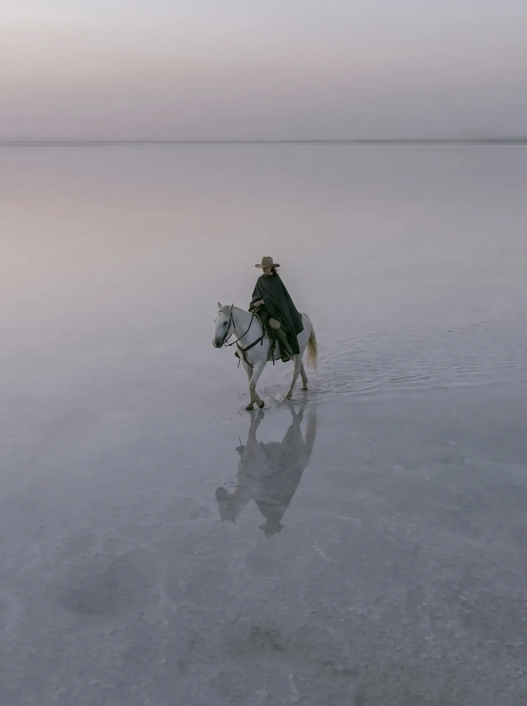 Cavalière en cape noire sur cheval blanc traversant un lac salé immense avec reflets au coucher du soleil rose