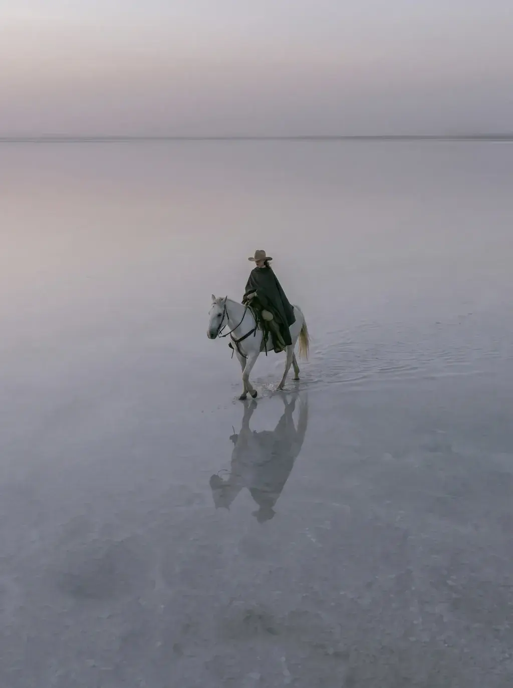 Cavalière en cape noire sur cheval blanc traversant un lac salé immense avec reflets au coucher du soleil rose