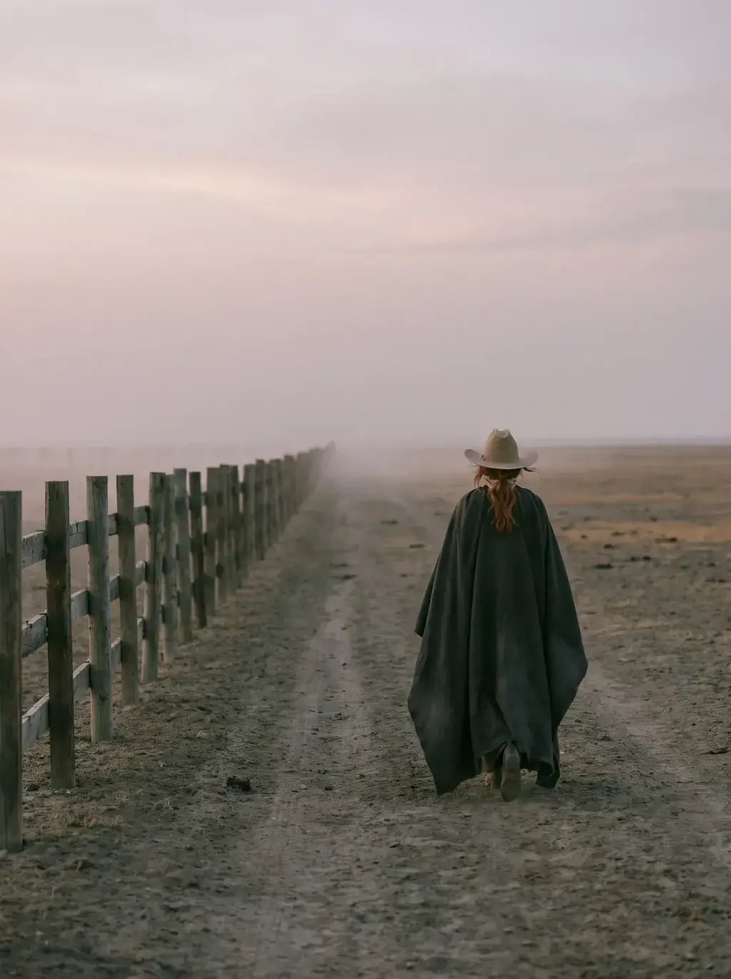 Cavalière en cape sombre de dos marchant le long d'une clôture de ranch vers la brume désertique