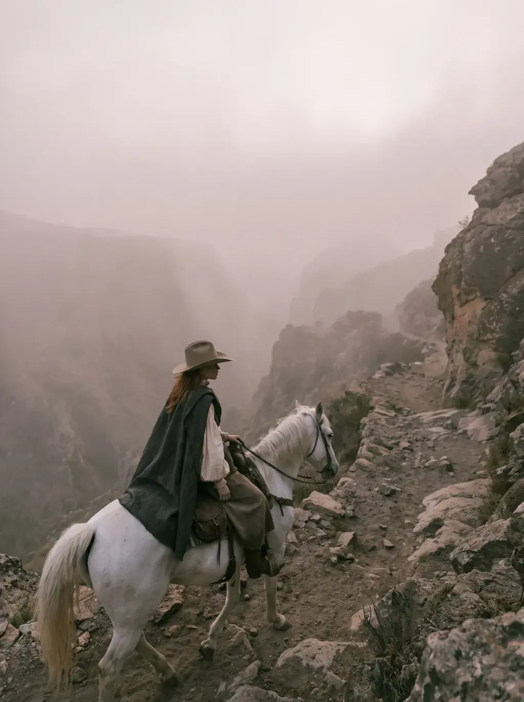 Cavalière en cape sombre sur cheval blanc sur un sentier de montagne escarpé dans la brume