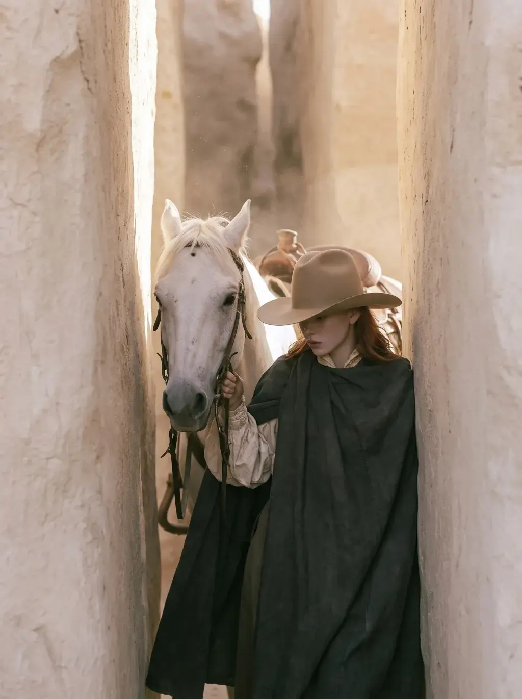 Cavalière rousse en cape noire guidant son cheval blanc dans un couloir étroit de murs en adobe