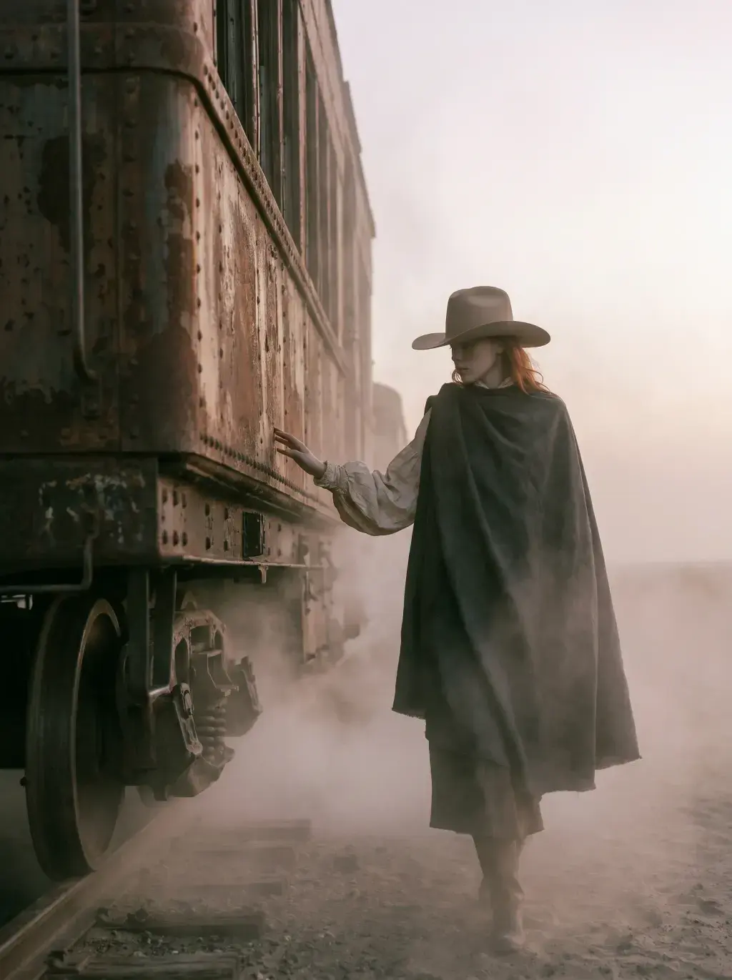 Cavalière rousse en cape sombre de profil touchant une locomotive rouillée abandonnée dans la brume