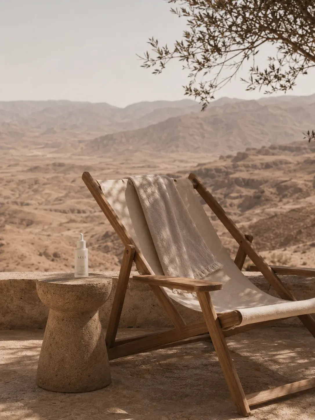 Chaise longue bois sur terrasse panoramique – table béton, flacon soin, vue désertique, olivier