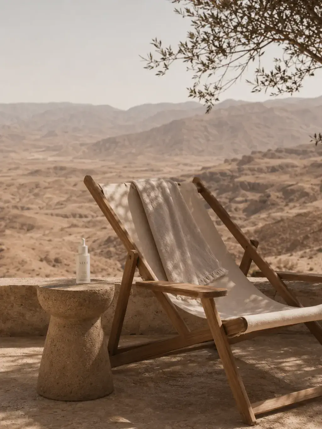Chaise longue bois sur terrasse panoramique – table béton, flacon soin, vue désertique, olivier