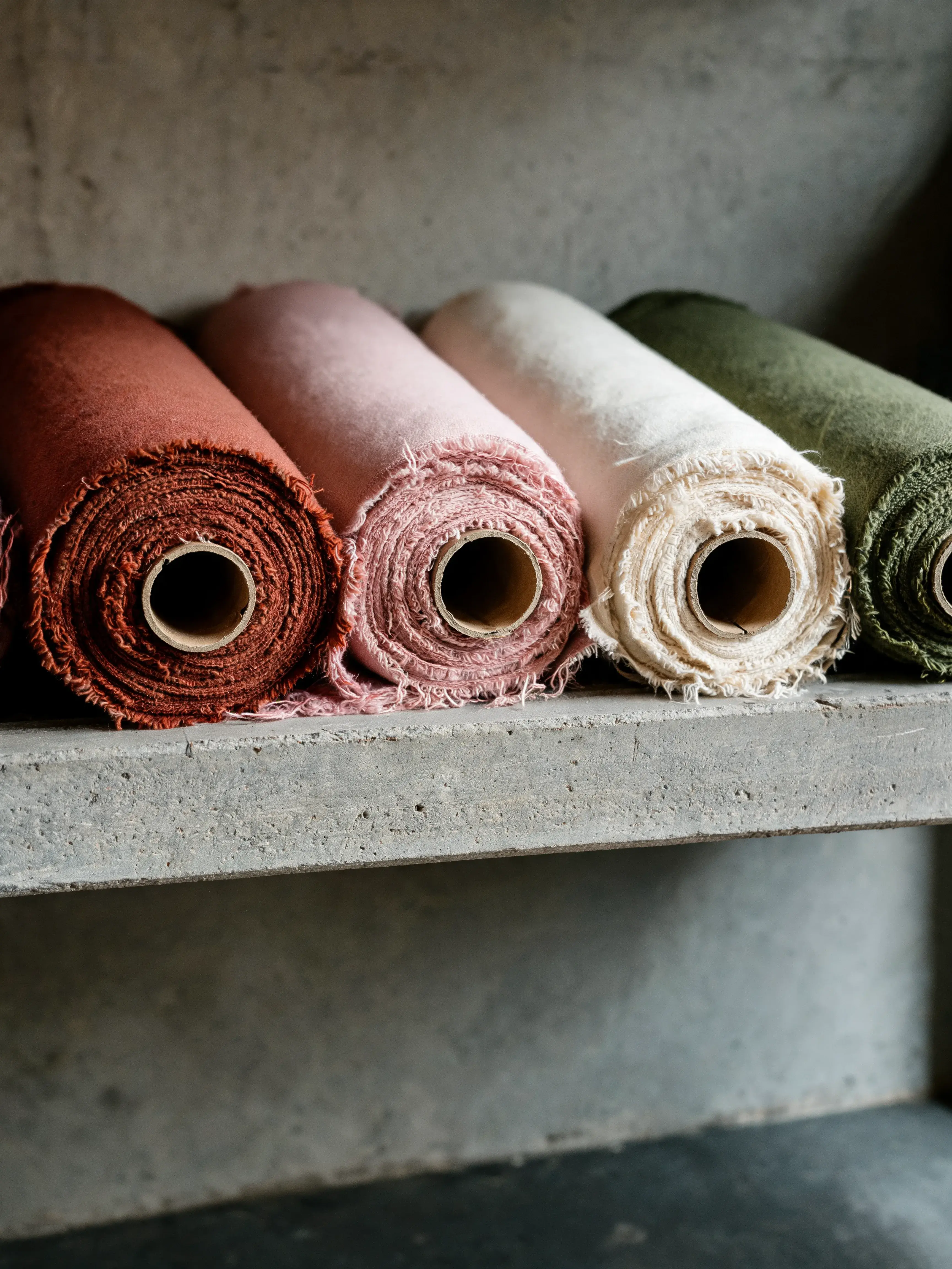 Cinq rouleaux tissu tons naturels alignés – terracotta rose crème vert, étagère béton, atelier sombre