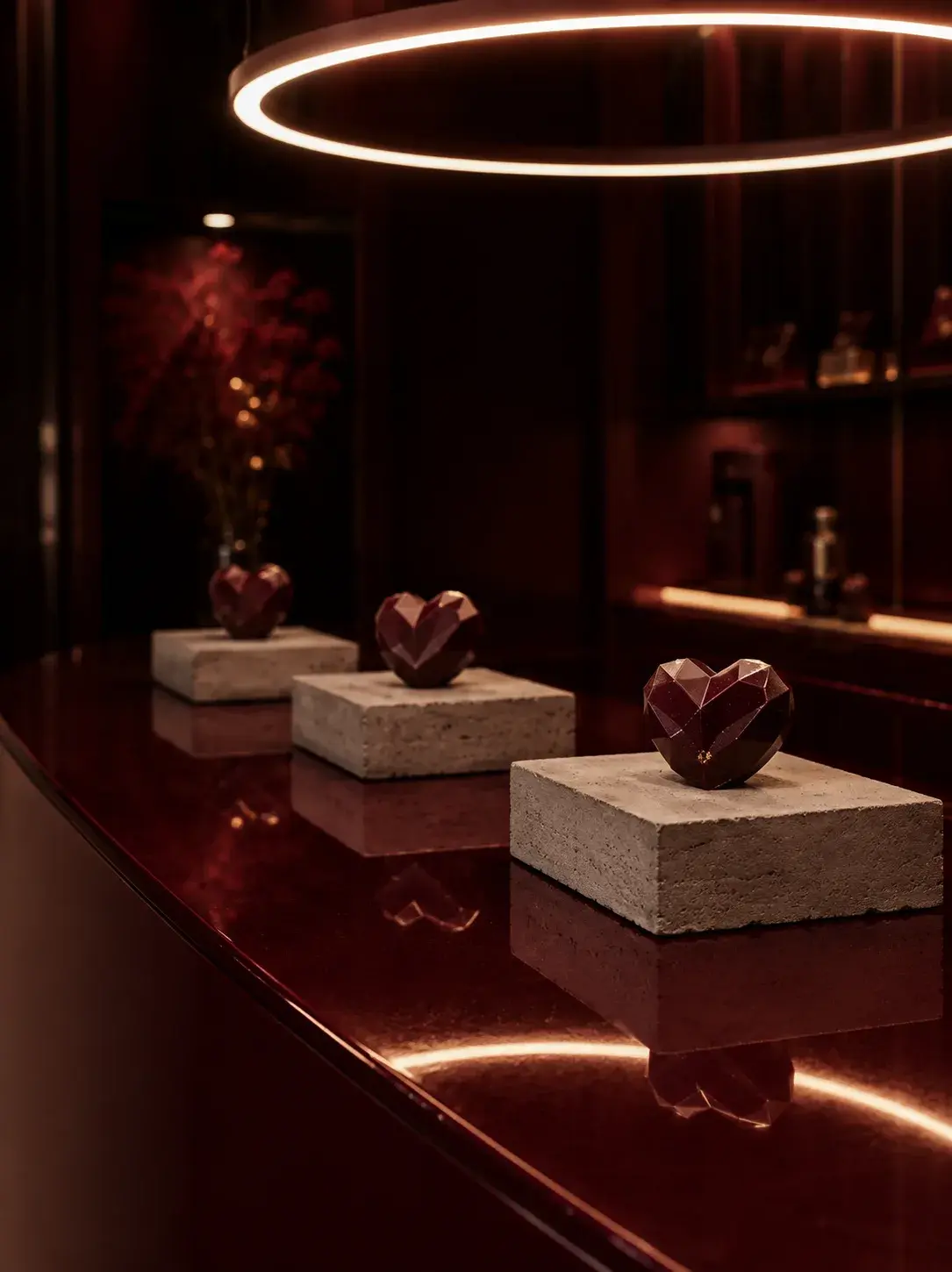 Comptoir rouge brillant avec trois cœurs en chocolat facettés sur socles pierre beige, lustre LED circulaire en arrière-plan