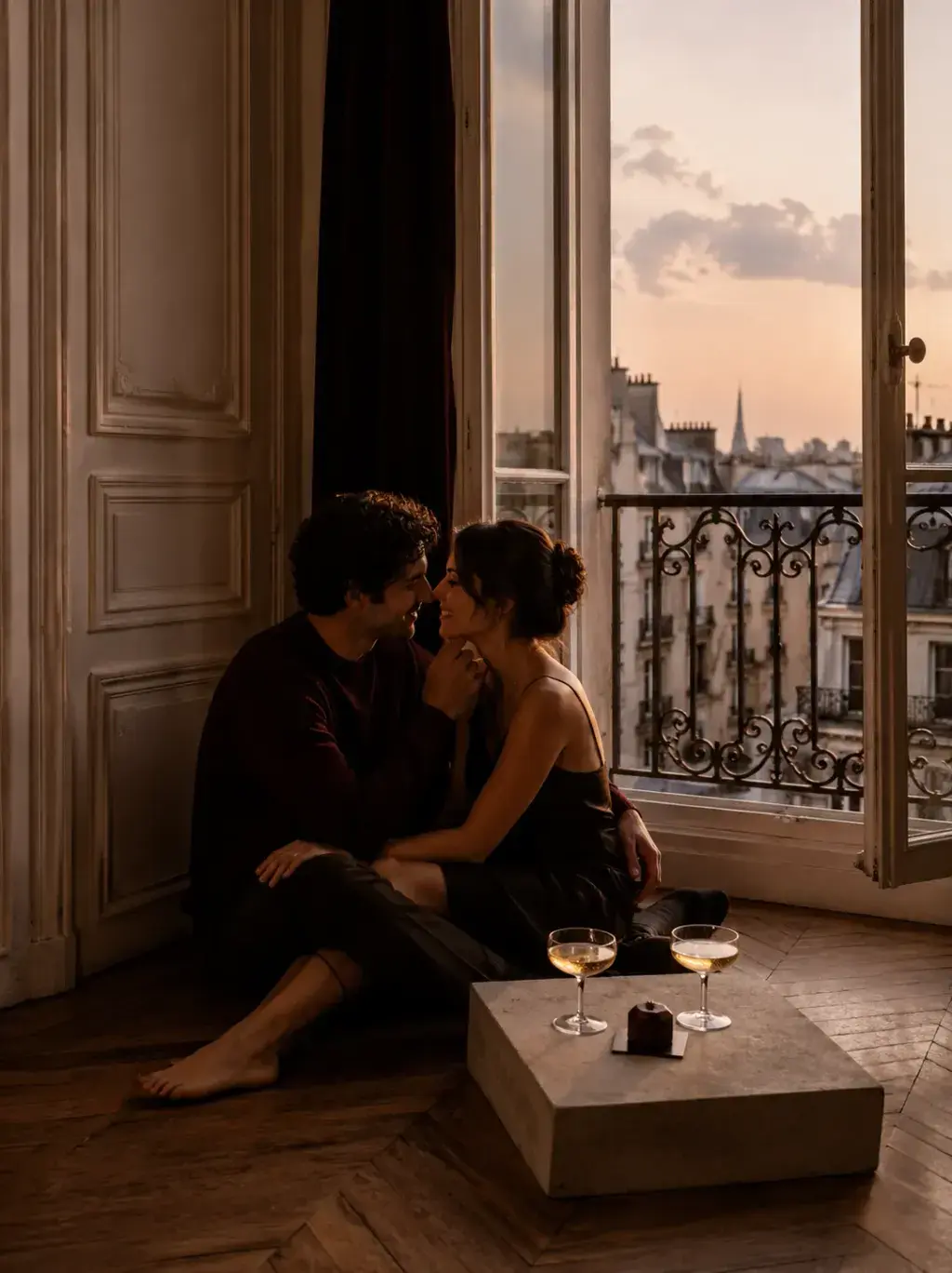 Couple parisien assis face à face devant fenêtre ouverte au coucher de soleil, verres de champagne sur table basse, bague de fiançailles