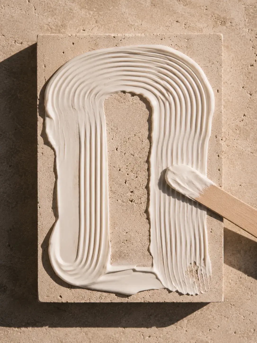 Crème blanche en motif courbe étalée avec spatule – bloc travertin, lumière rasante, vue de dessus
