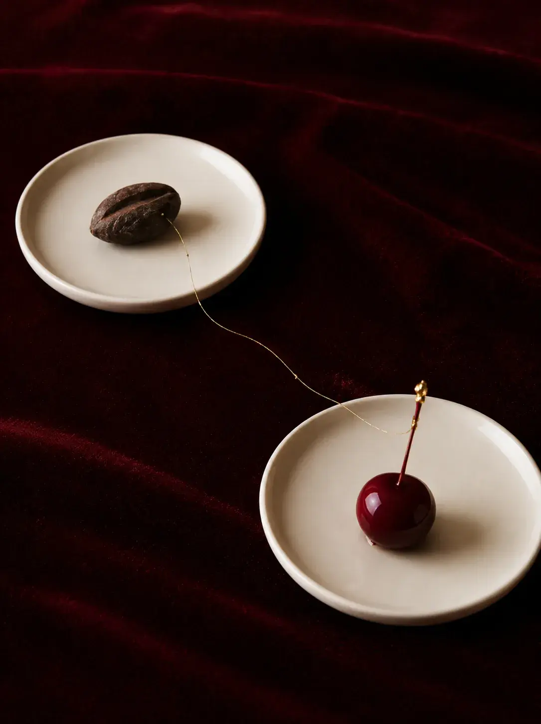 Deux assiettes blanches sur velours bordeaux – grain de cacao et cerise reliés par fil doré