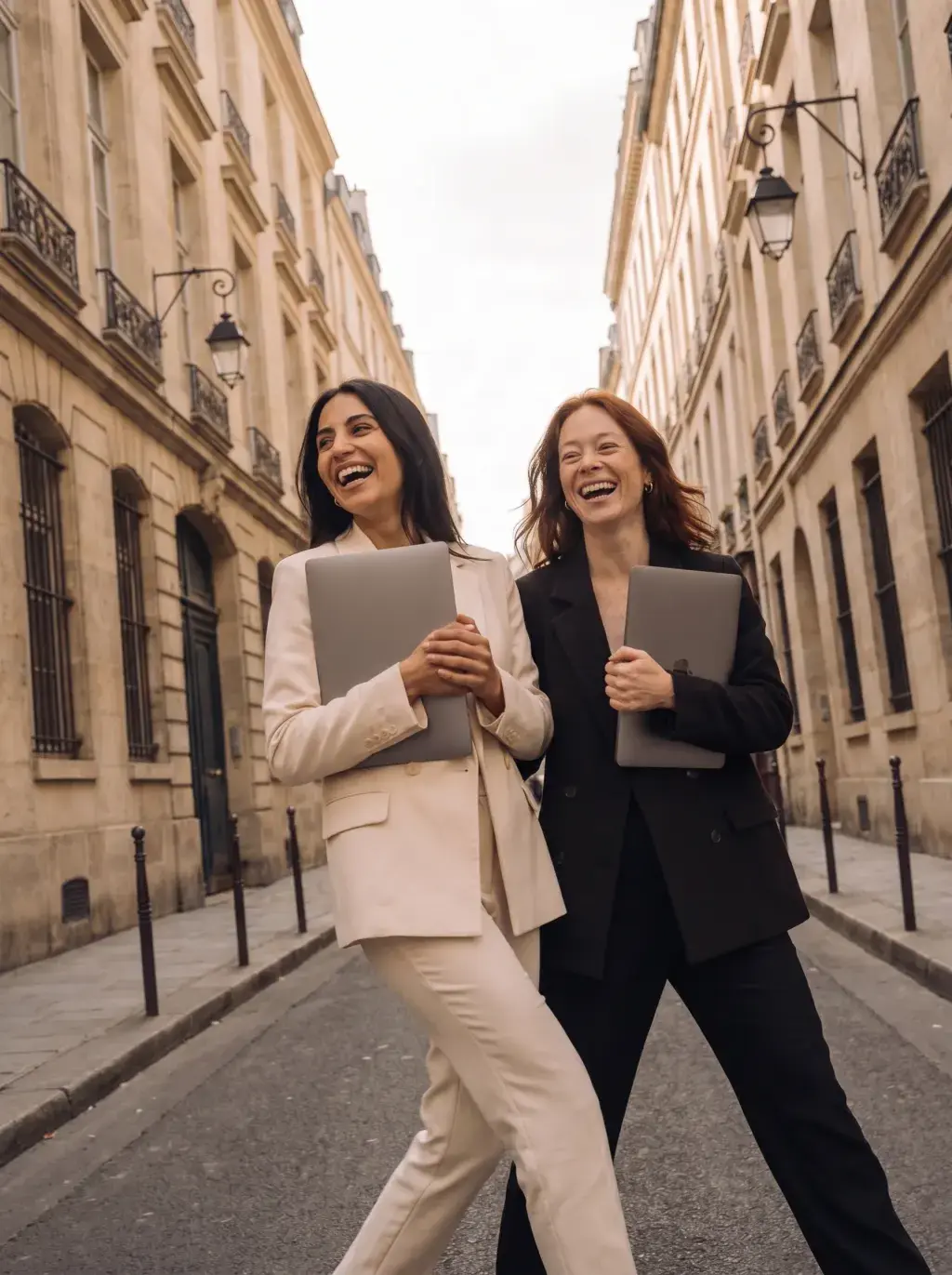 Deux associées marchant en riant dans une rue haussmannienne avec leurs laptops