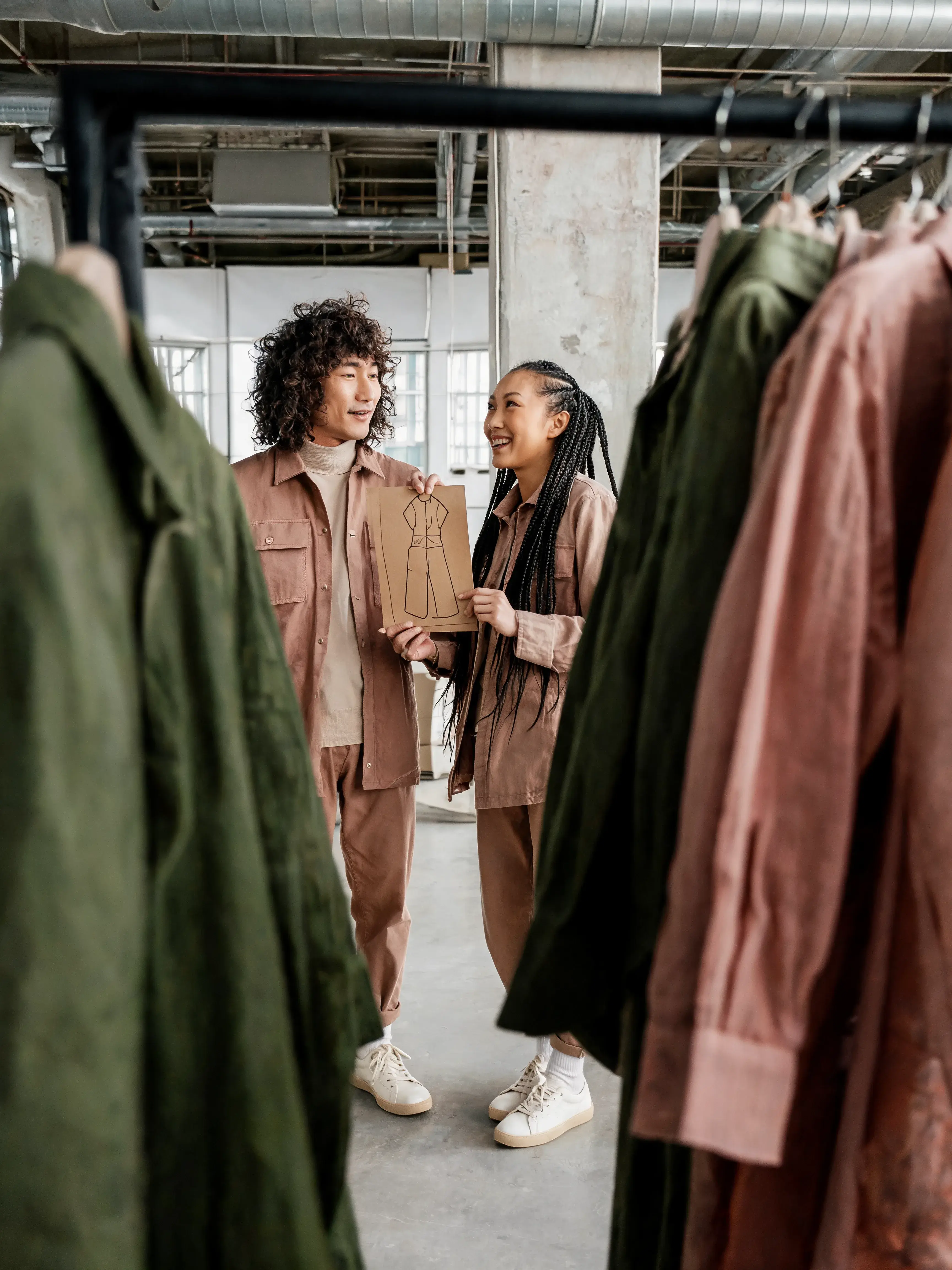 Deux collègues conversant entre portants mode – croquis silhouette carton, showroom industriel