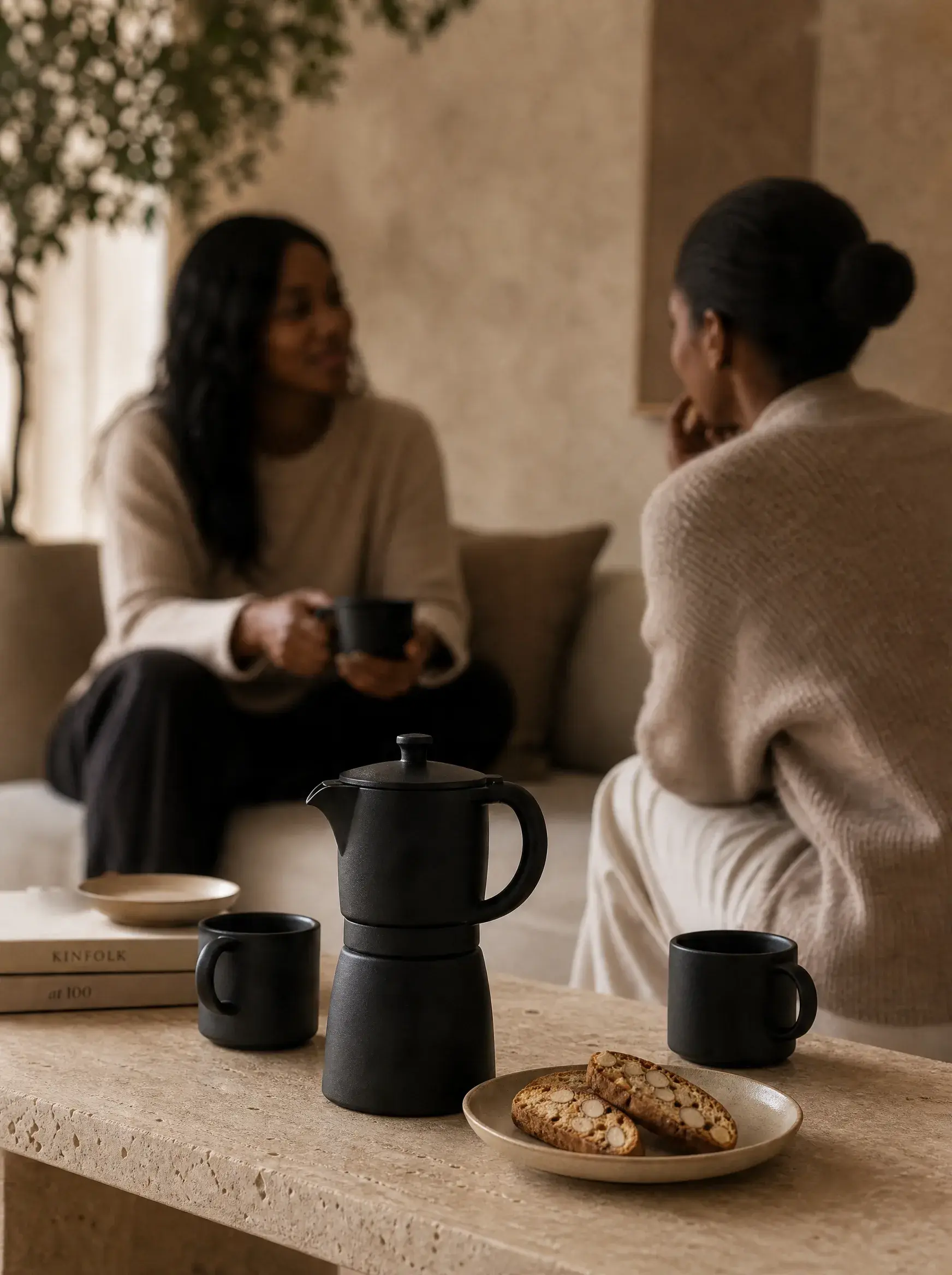 Deux femmes discutant avec mugs café – canapé, cafetière moka, biscotti, table travertin, plante