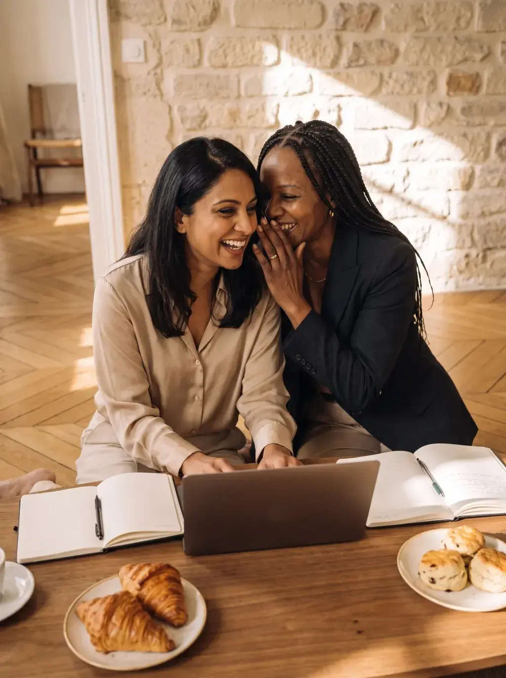 Deux femmes riant ensemble autour d'un laptop avec croissants dans un intérieur en pierre