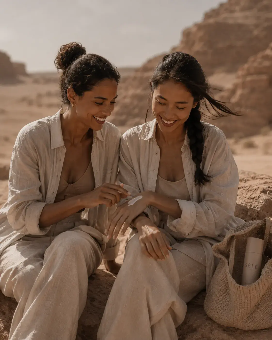 Deux femmes souriantes sur rocher désertique – application de soin, tenues lin, sac jute, complicité