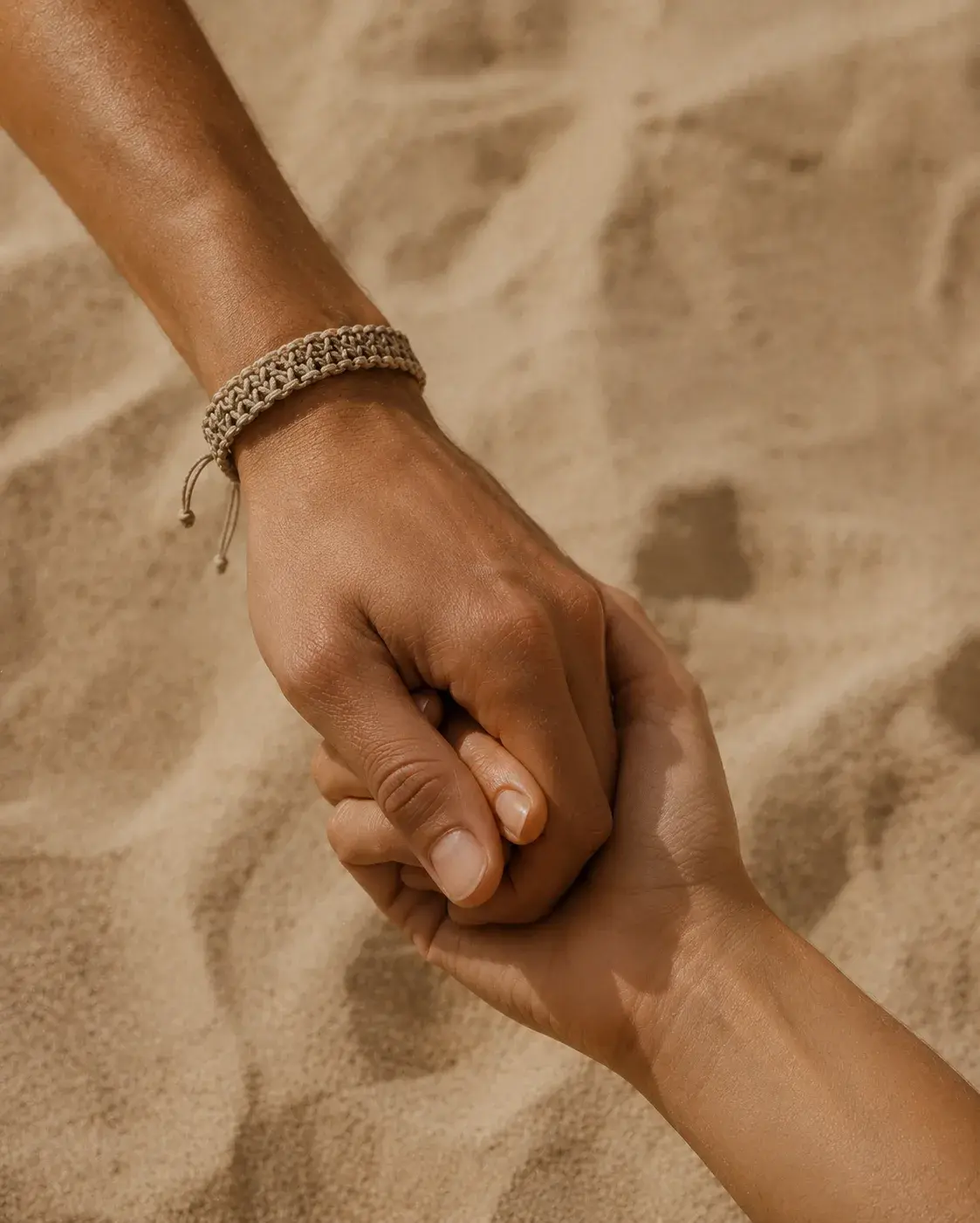 Deux mains se tenant sur fond de sable – bracelet macramé beige, diversité, peau noire et peau claire