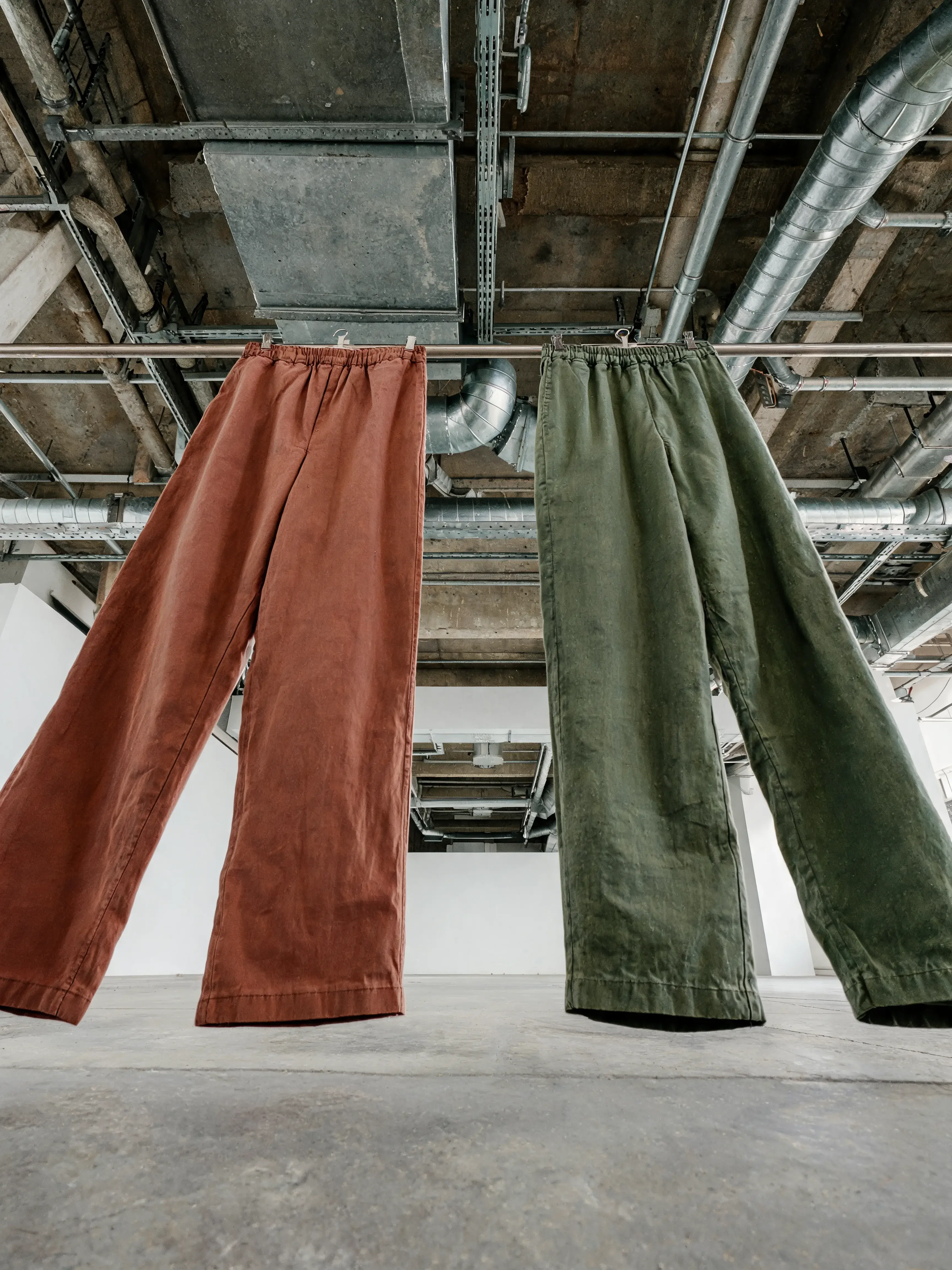 Deux pantalons larges suspendus – terracotta et vert, atelier industriel, plafond béton
