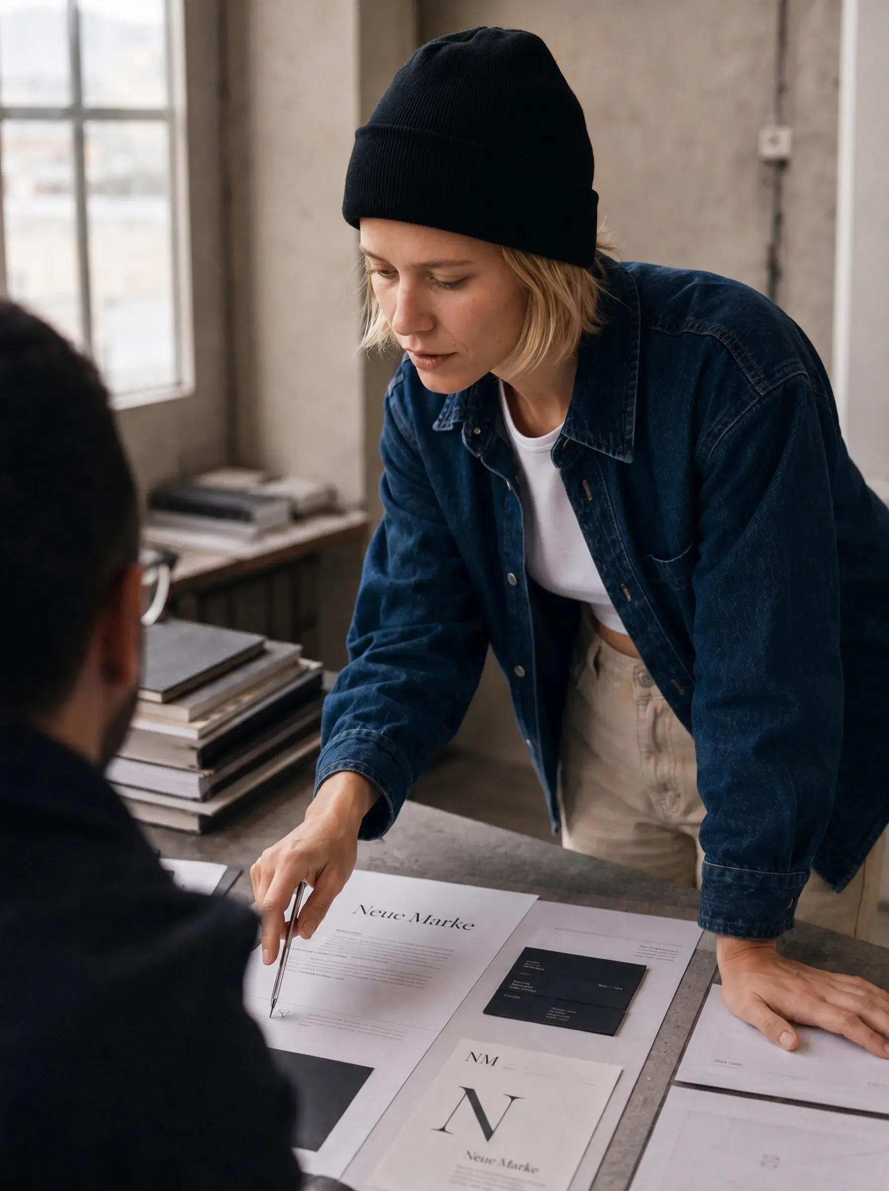 Directrice créative présentant l'identité visuelle « Neue Marke / NM » à un client devant documents étalés sur table