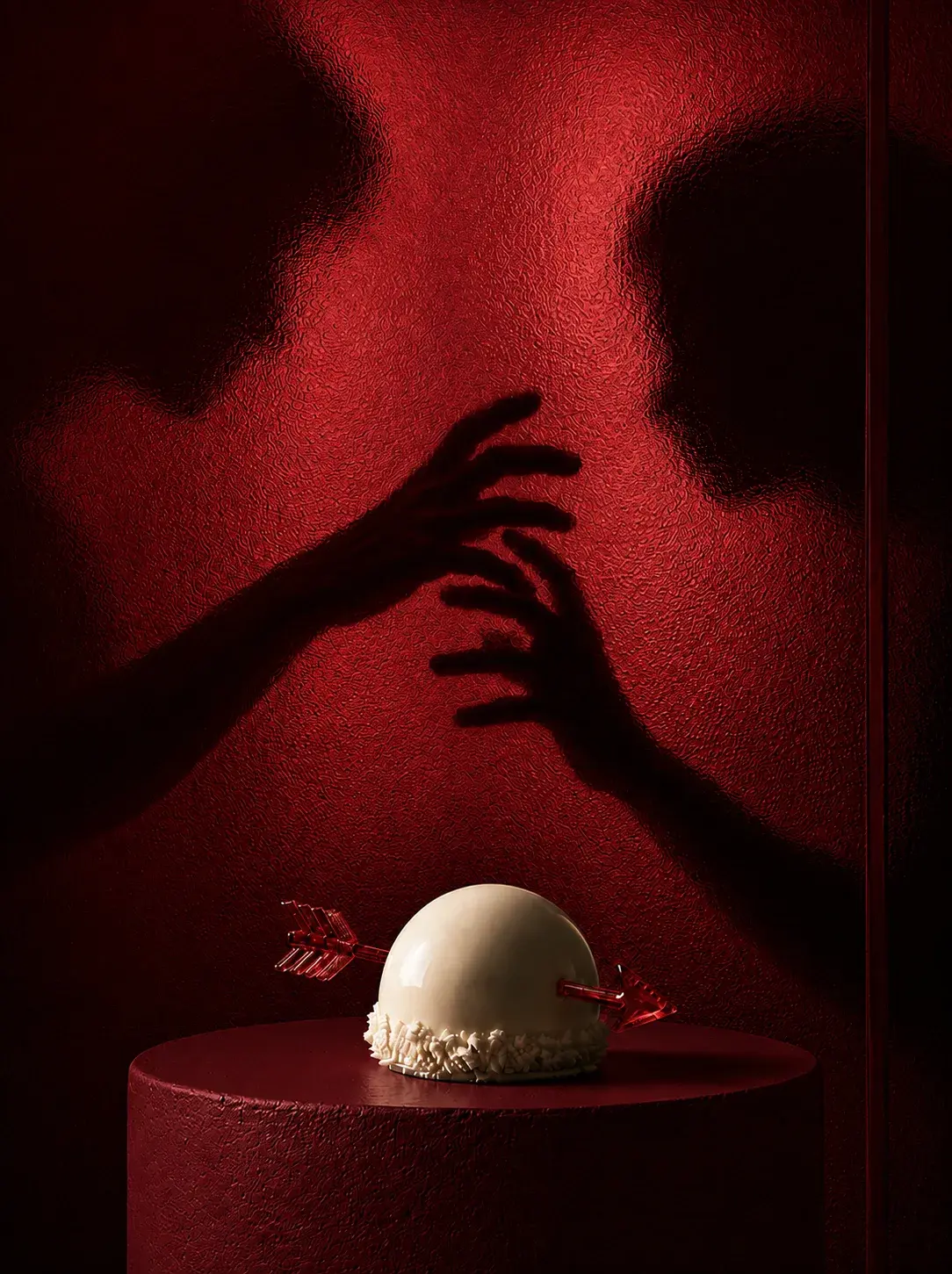 Dôme pâtissier blanc avec flèche de cupidon sur socle velours rouge, deux ombres de mains se rejoignant sur mur rouge texturé