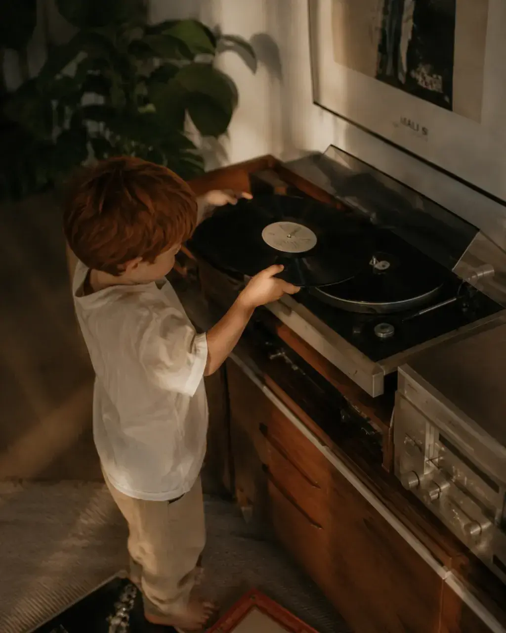 Enfant roux de dos actionnant une platine vinyle – salon, lumière chaude et plante verte