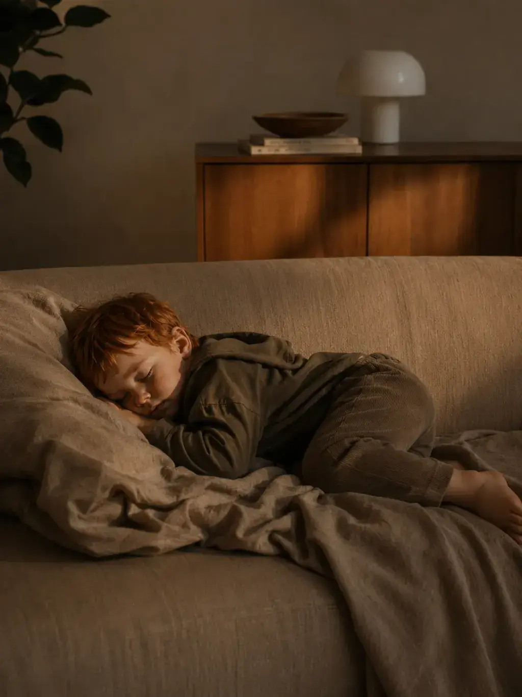 Enfant roux endormi sur un canapé beige – lumière douce du soir, intérieur cosy
